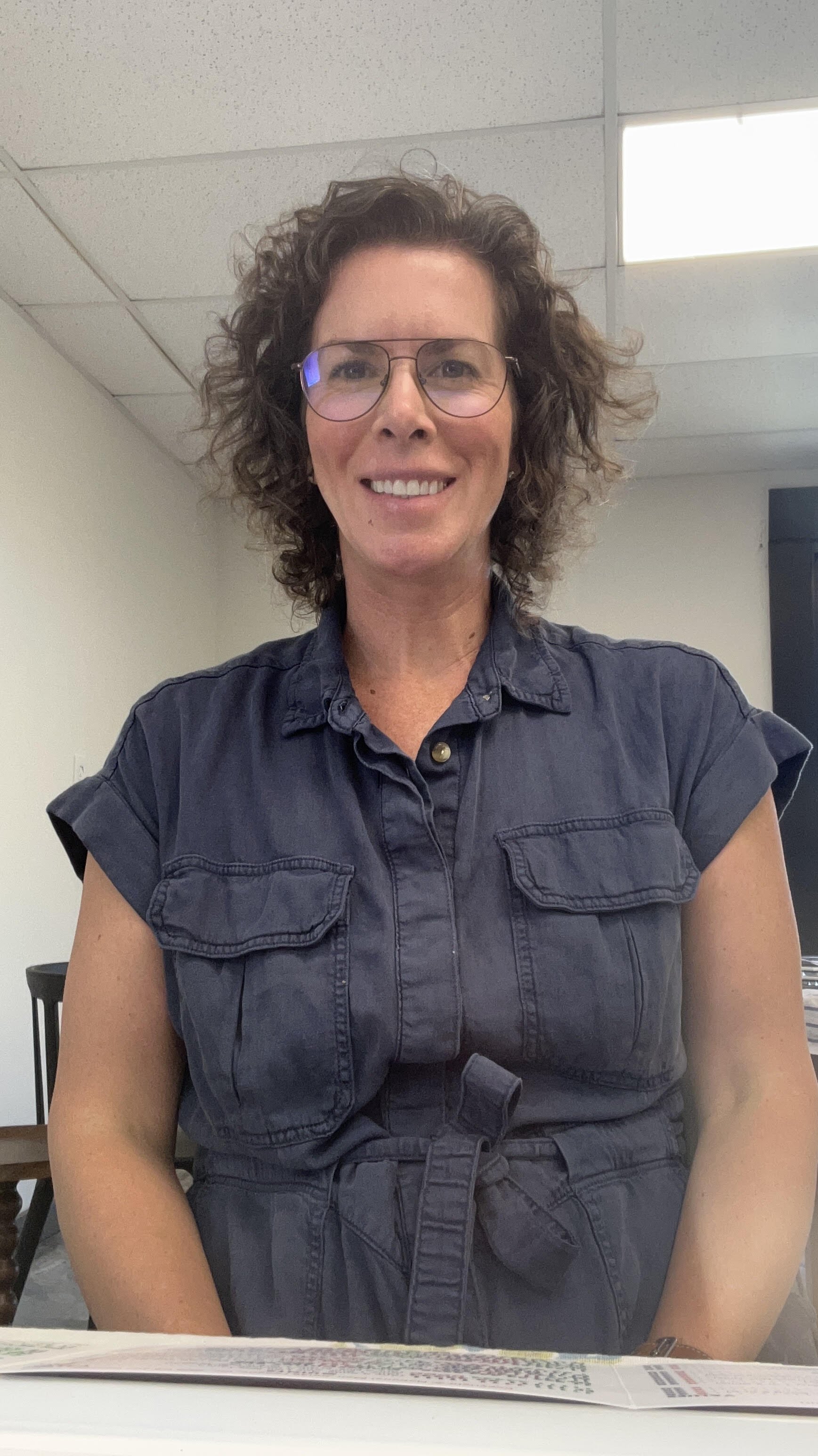 A woman with glasses and curly hair, wearing a sleeveless denim button-up shirt with a tied waist, sitting at a table in an office setting with a ceiling of white tiles and overhead lights.