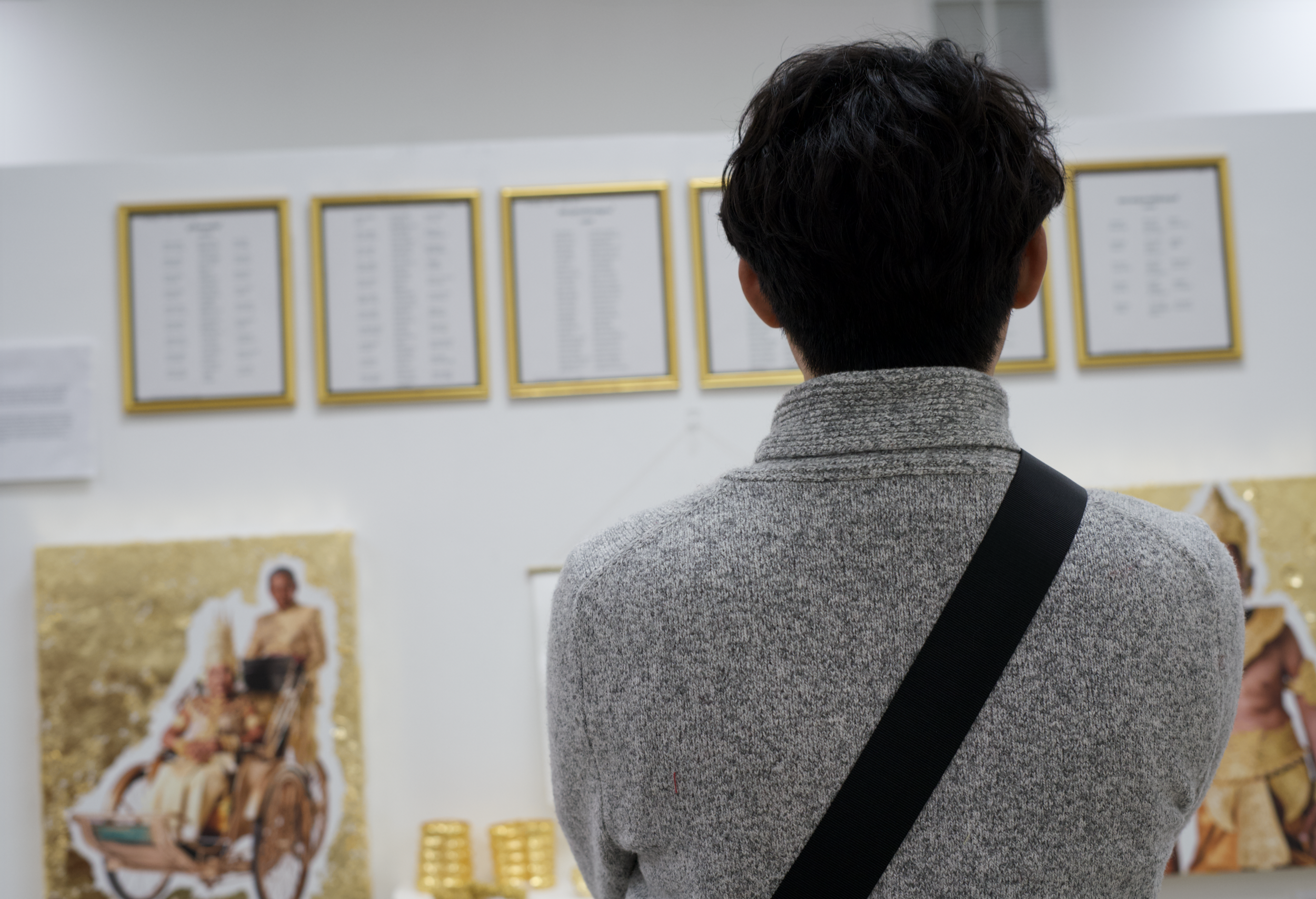 A person with short dark hair and a gray sweater stands with their back to the camera, looking at posters on a wall in an art gallery or museum.