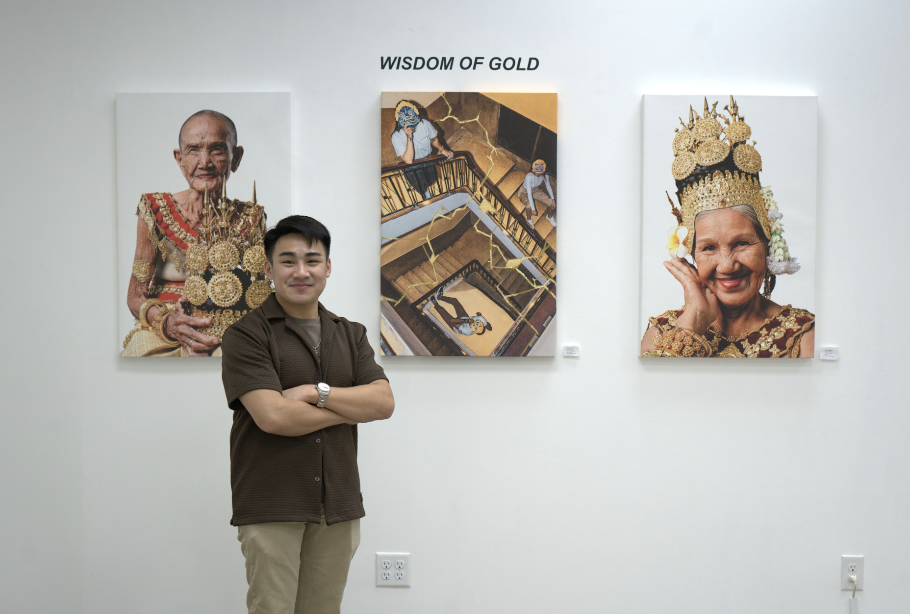 A man standing with his arms crossed in front of three large portraits on a white gallery wall. The portraits depict a woman in traditional Thai attire, including ornate gold jewelry and headdresses, with the middle portrait showing a scene of children on a staircase. The gallery has a clean white background and visible electrical outlets near the floor.