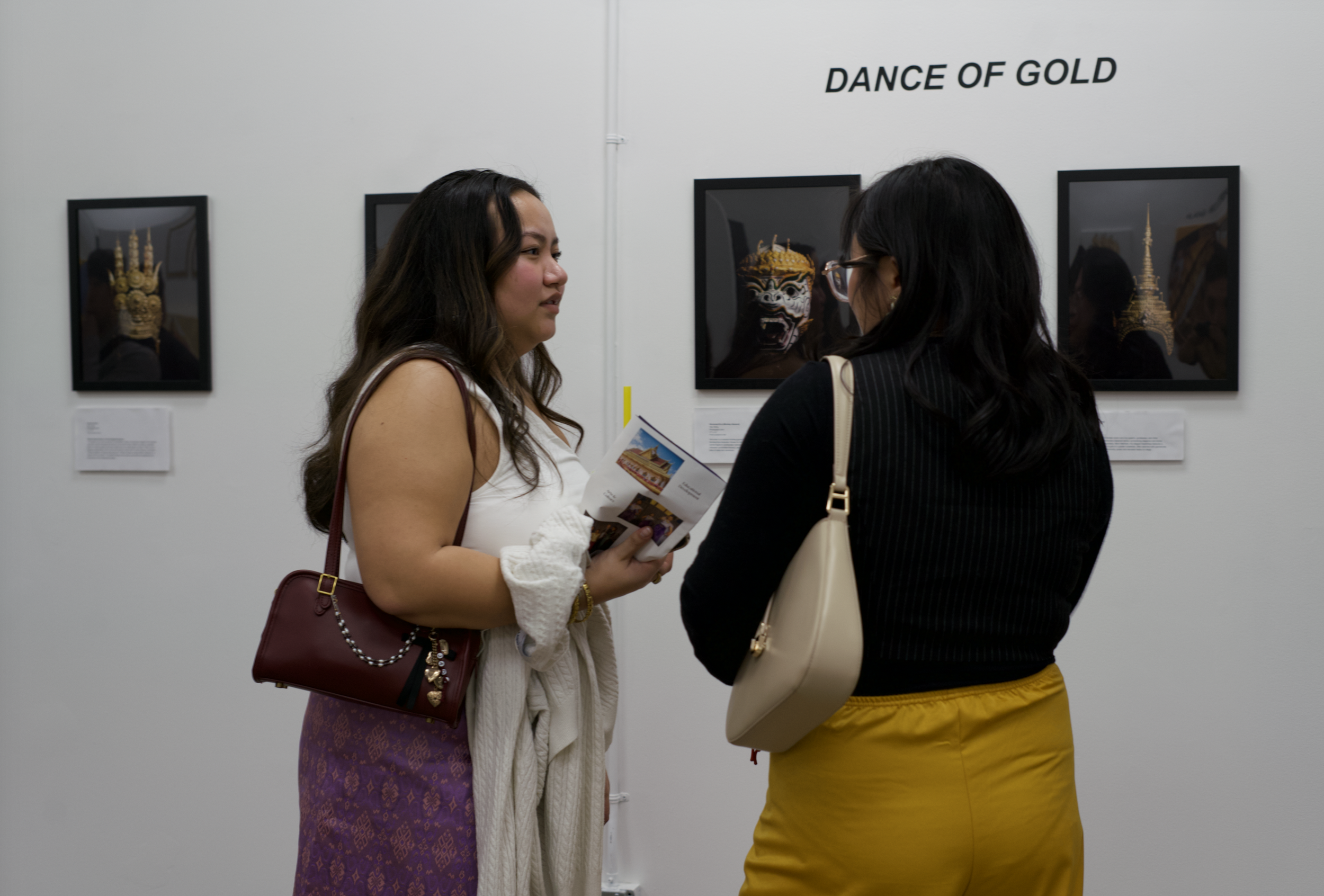 Two women are talking in an art gallery with framed artwork on the wall behind them. One woman is holding a brochure, and the other is wearing glasses and a beige purse.