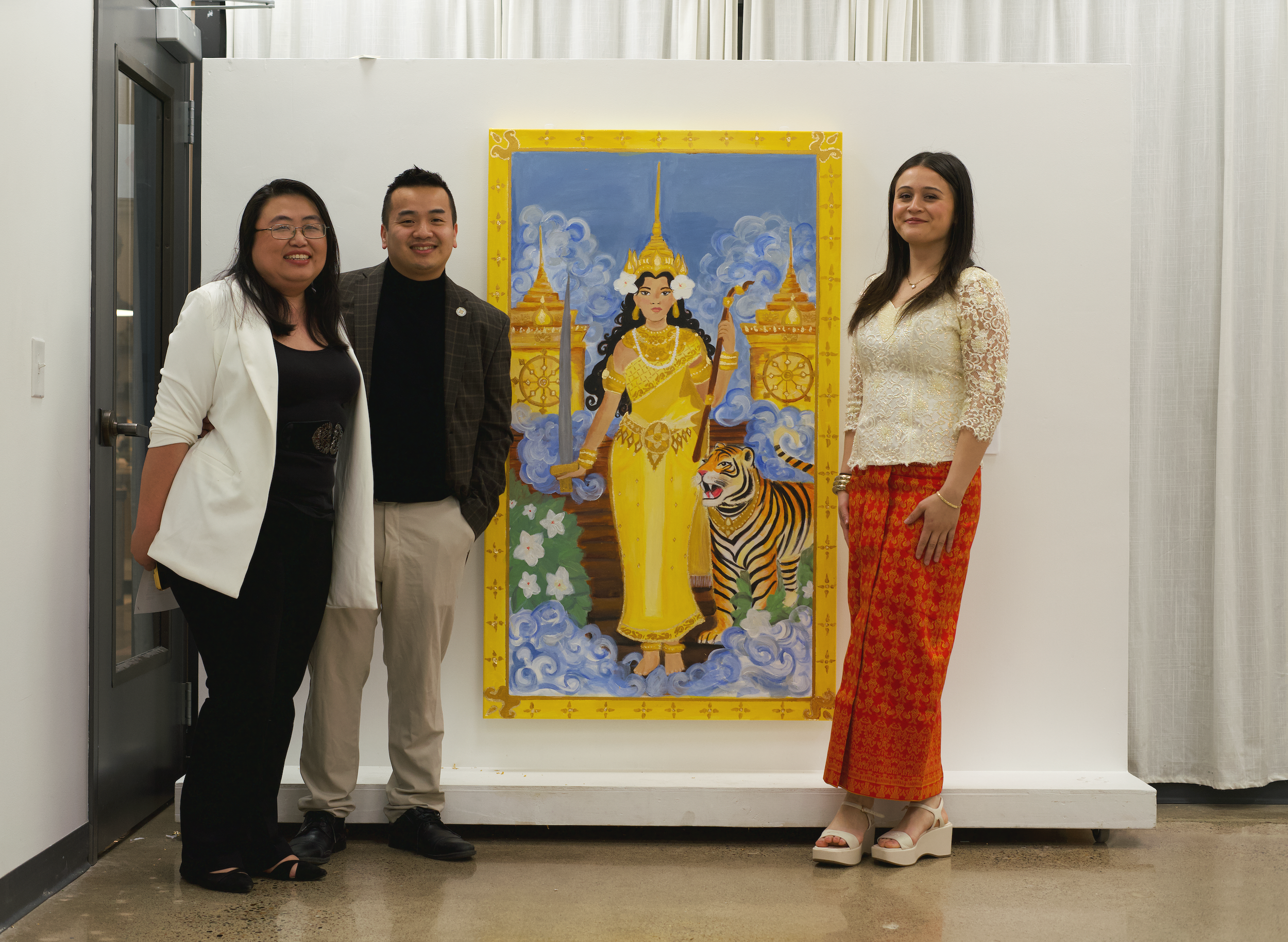 Three people standing in front of a colorful painting of a regal woman in traditional Thai attire with a tiger at her feet. The group consists of two women and one man, all dressed semi-formally, smiling for the photo in what appears to be an indoor 