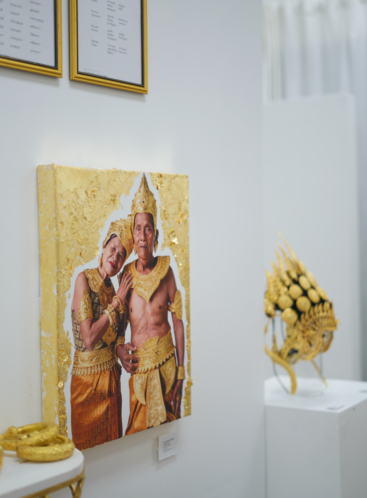 Gallery display featuring a portrait of elderly Southeast Asian couple dressed in traditional gold and orange ceremonial attire, and an ornate gold headdress sculpture on a stand.