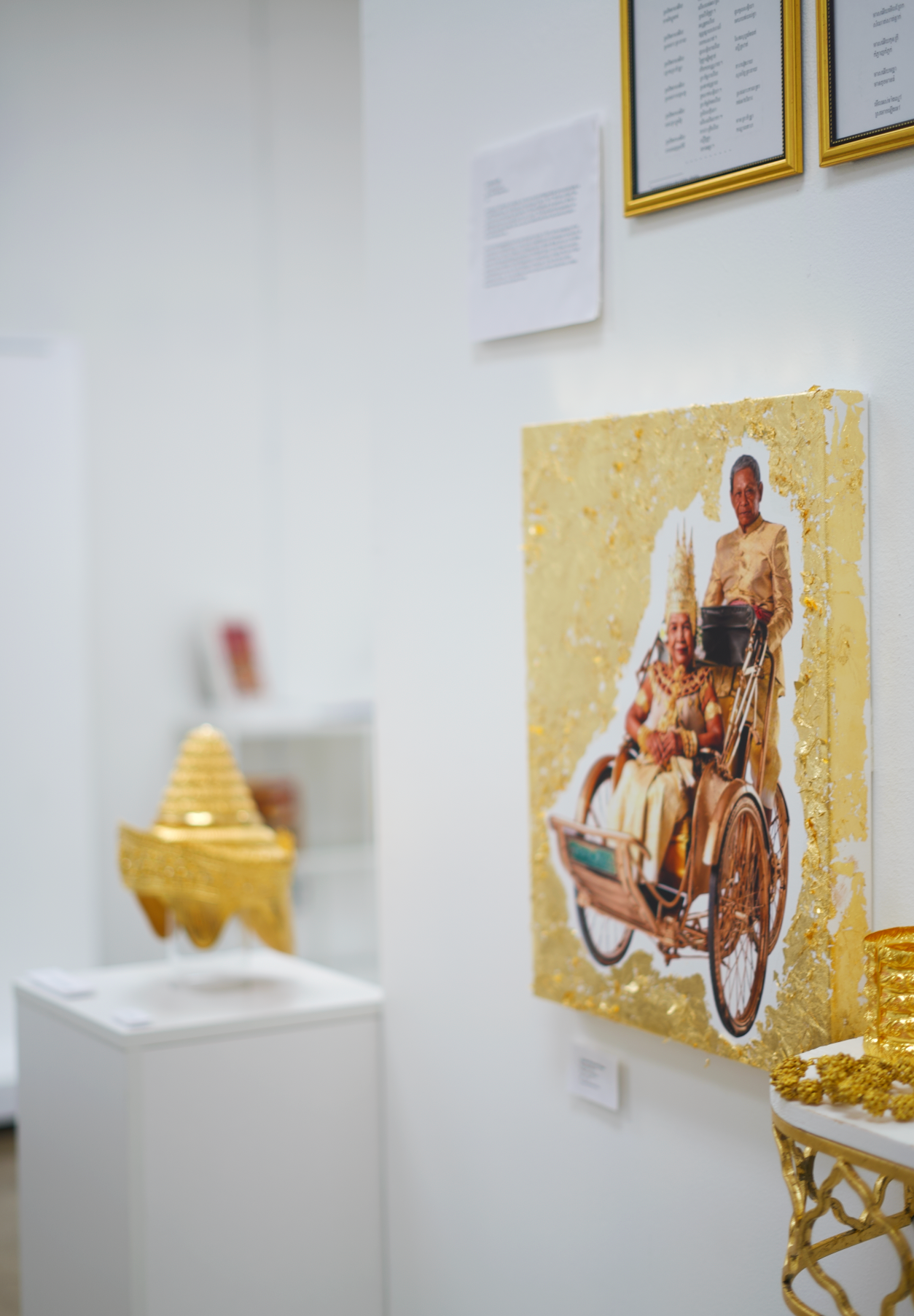 A gallery wall with framed texts, a colorful painting of women, and a golden sculpture of a temple, displayed in an art exhibition setting.