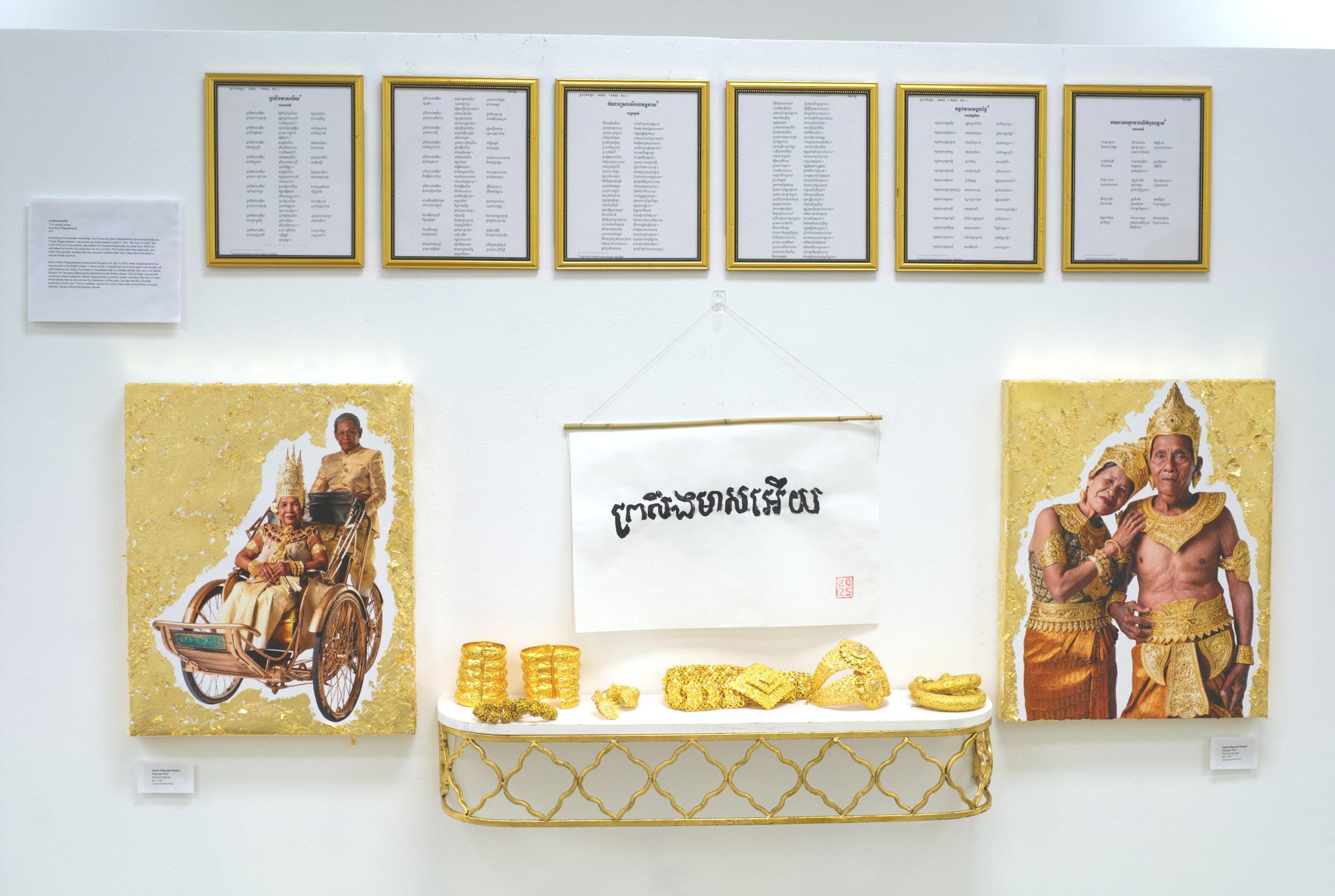 Exhibition display featuring posters of elderly people in traditional and royal attire, gold jewelry, and accessories, with framed documents and gold decorative items on a white wall.