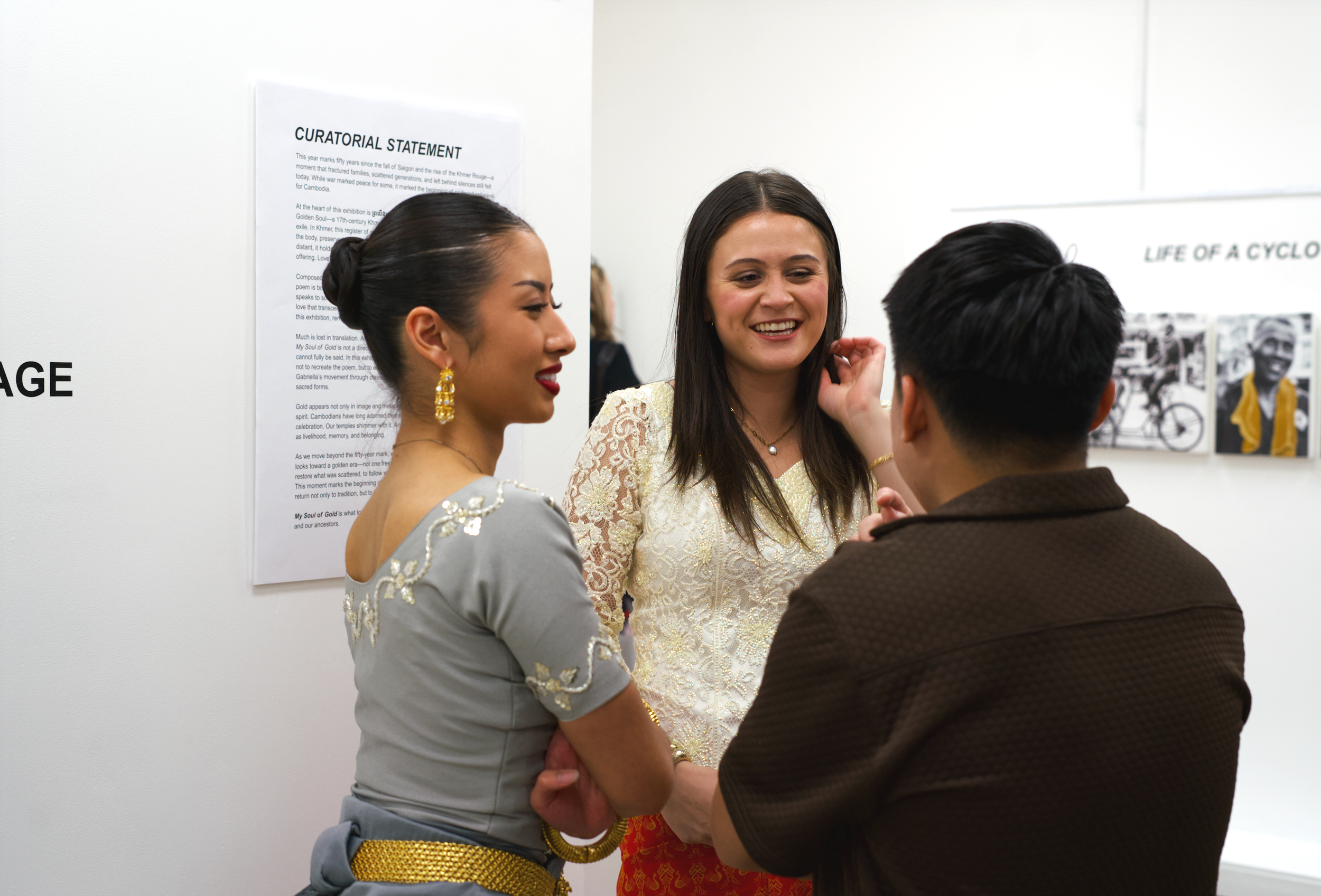 Three people, two women and one man, are engaged in conversation at an art exhibition, standing near a white wall with a curator statement and artist photographs, smiling and touching each other's faces.
