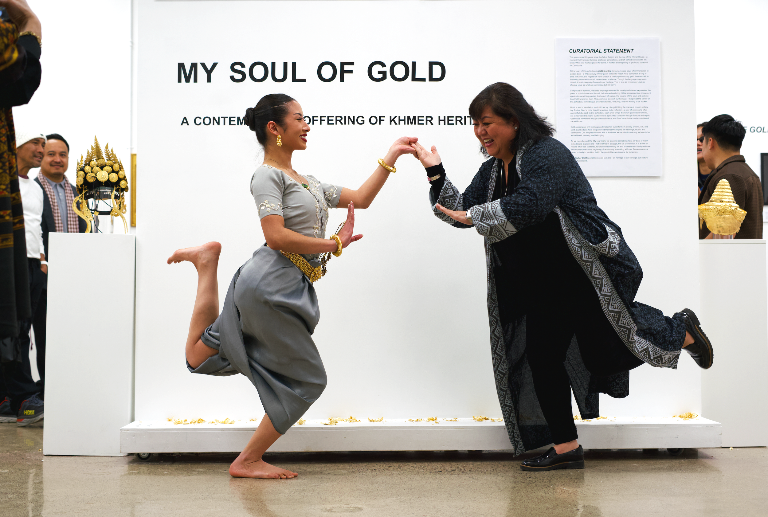 A woman in traditional Khmer attire kneeling on one knee and smiling, holding hands with an older woman who is also smiling and wearing black clothing, in front of a large sign that reads "MY SOUL OF GOLD" with the subtitle "A CONTEMPORARY OFFERING O