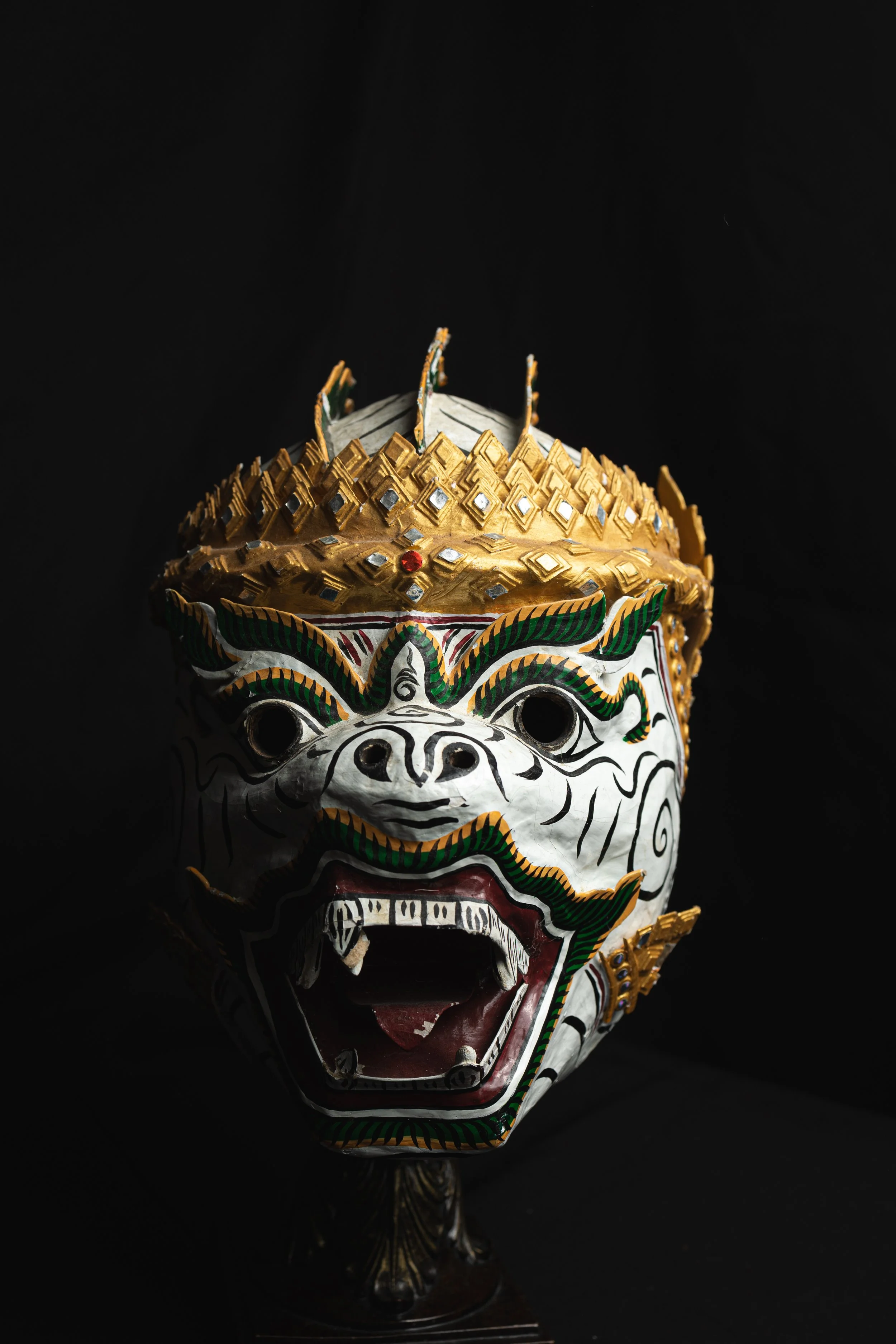Colorful, detailed mask with a fierce expression, decorated with gold, green, and red accents, and mounted on a stand against a black background.