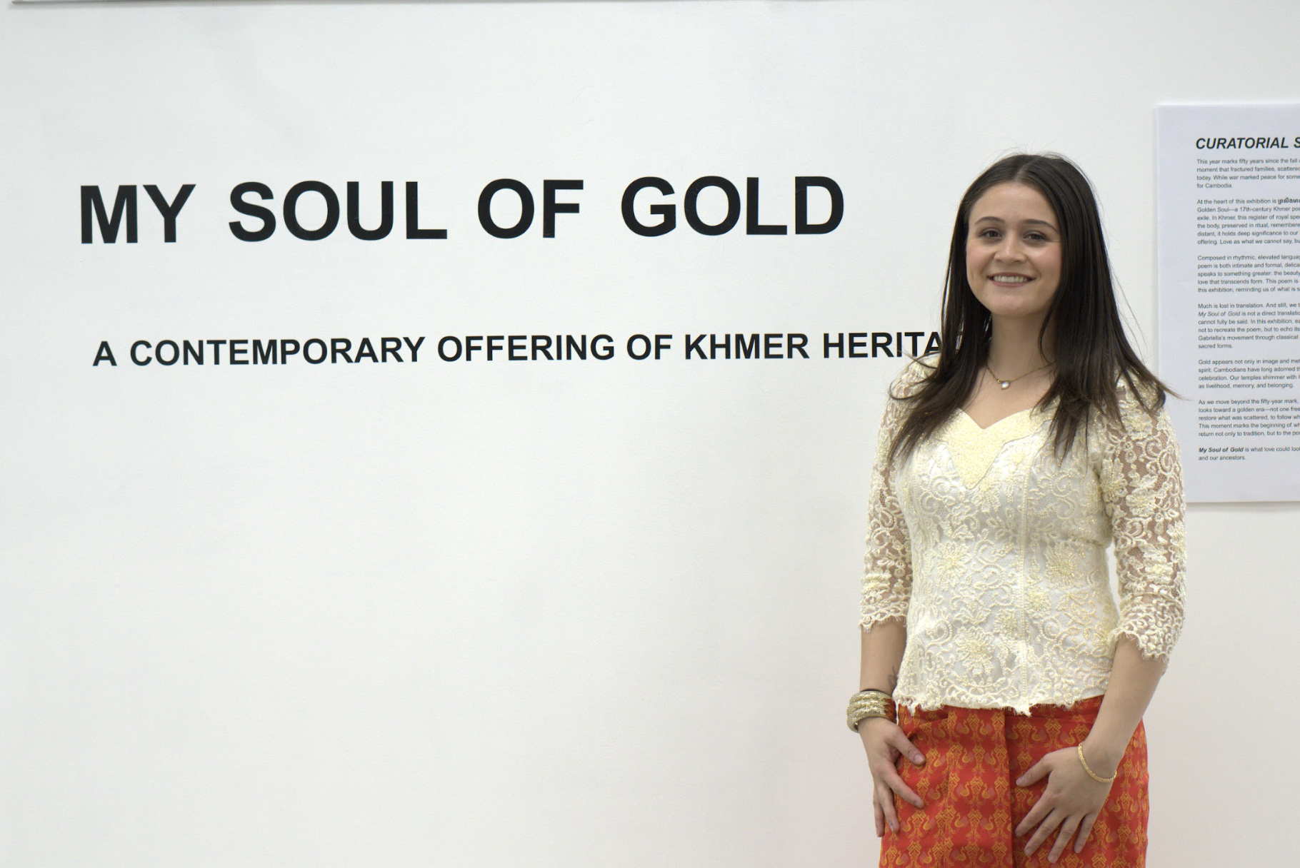 A woman standing in front of a gallery wall with a white background, wearing a cream lace top and red patterned pants, smiling at the camera.