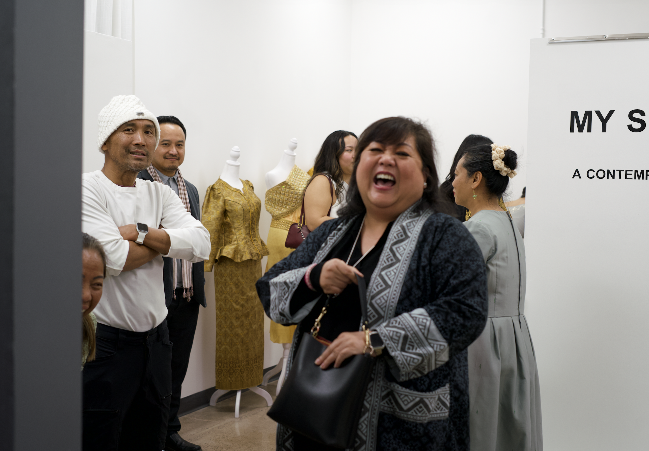 People attending an art exhibition or gallery, with mannequins dressed in traditional Asian clothing and a woman laughing in the foreground.