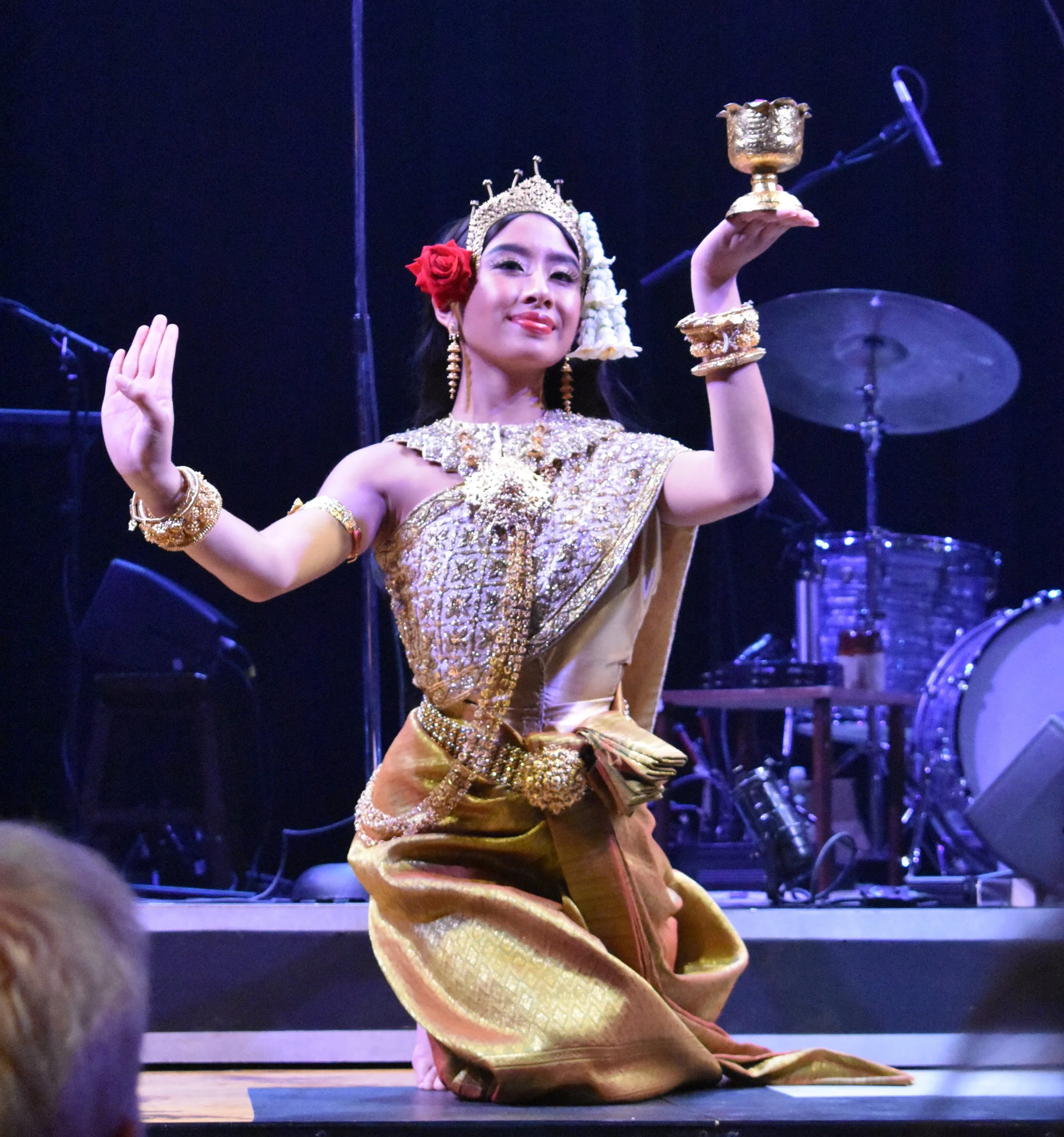 A woman dressed in traditional Thai attire is sitting on the stage, holding a golden chalice in her right hand, with a confident expression. She wears gold jewelry, a gold headdress decorated with flowers, and a gold and pink outfit. The background features stage lighting and musical equipment.