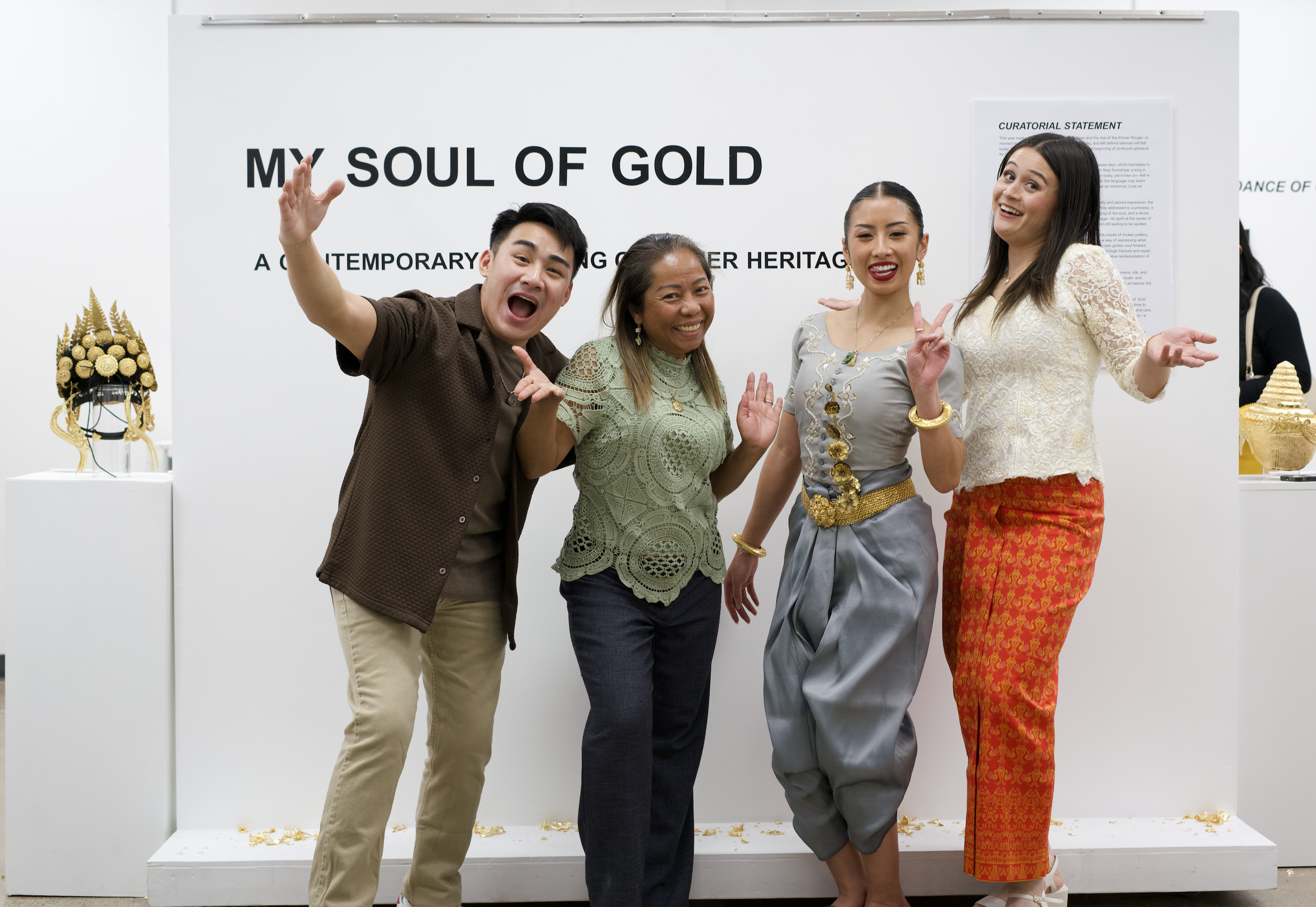 Four people, two men and two women, standing together in front of a white display board that reads "MY SOUL OF GOLD." They are smiling and posing cheerfully, with the woman on the right gesturing with her hand. The display area includes golden artifa
