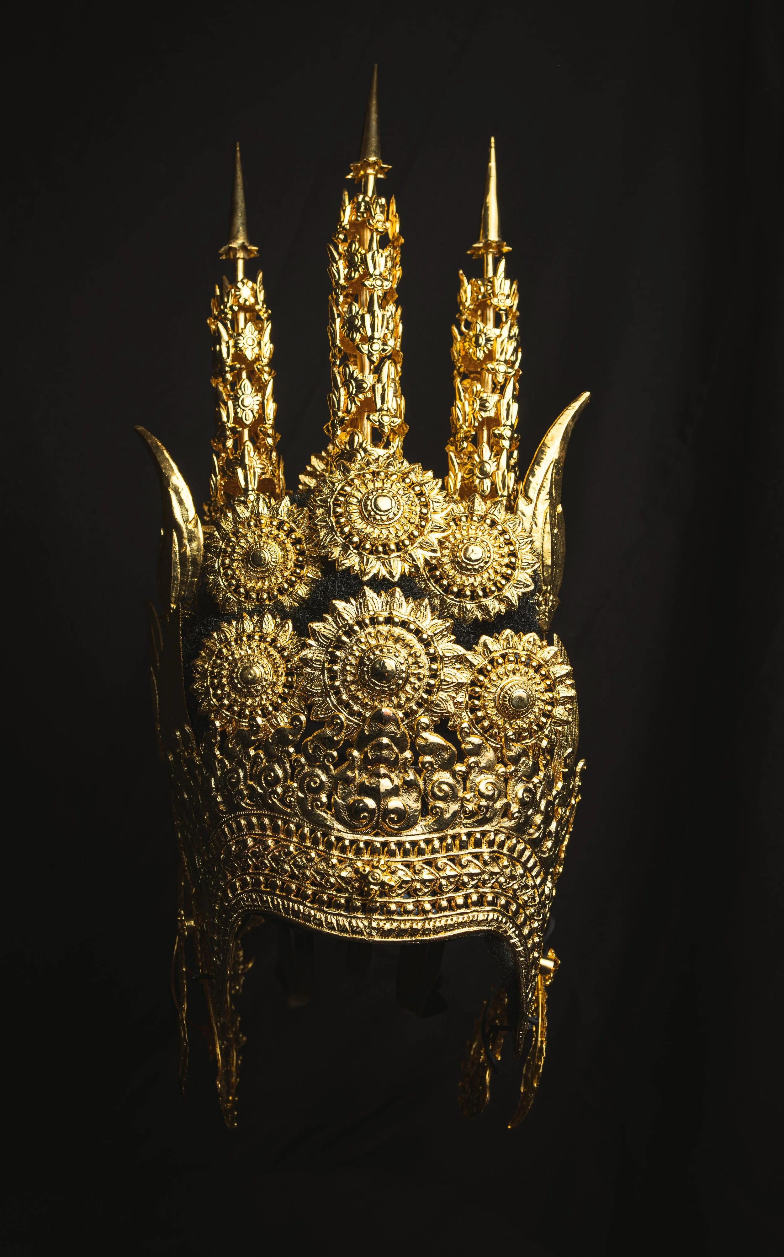 An ornate gold headpiece or crown with intricate floral and sunburst designs, set against a black background.