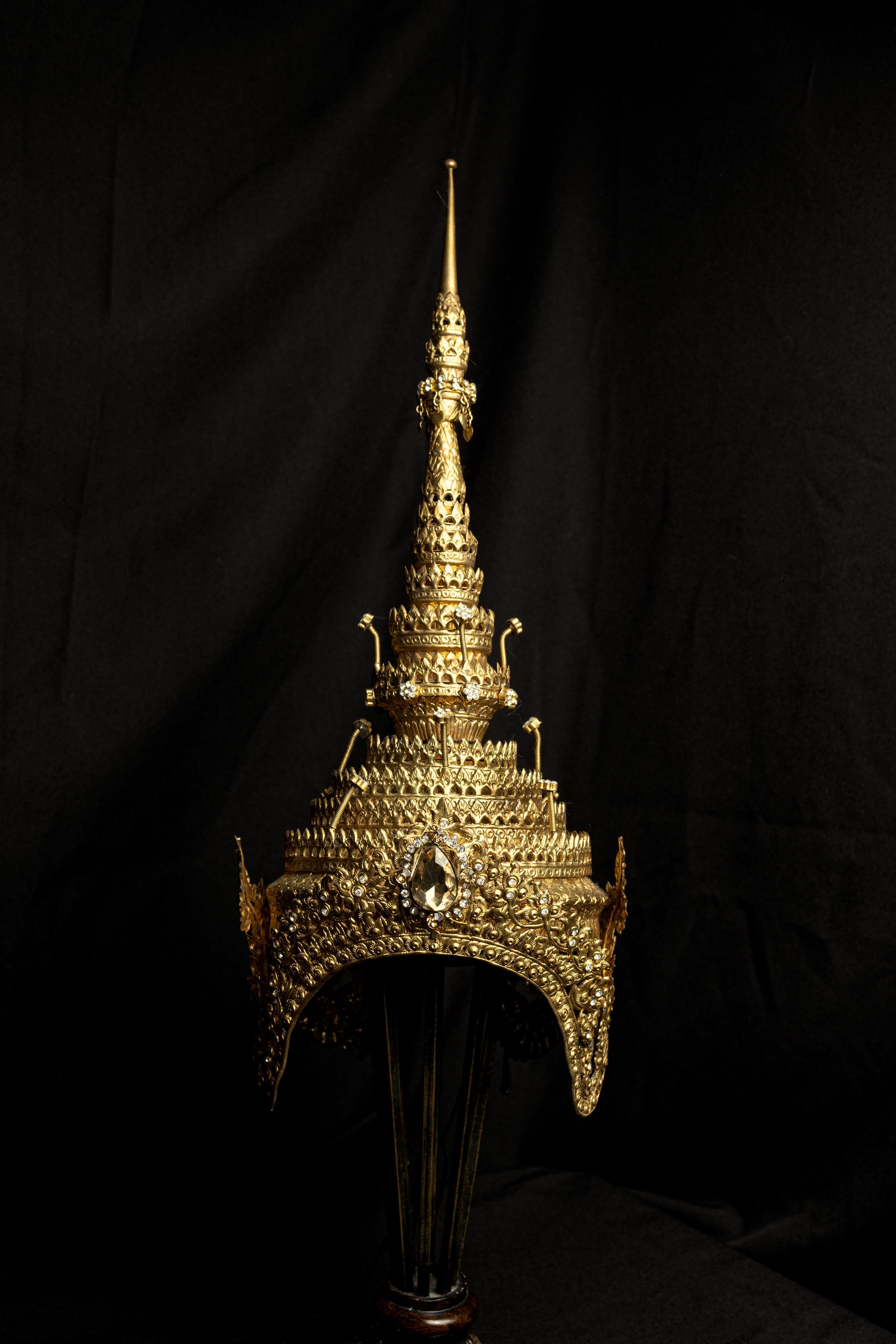 A detailed, ornate golden crown with intricate designs, gemstones, and a pointed spire at the top, set against a dark background.