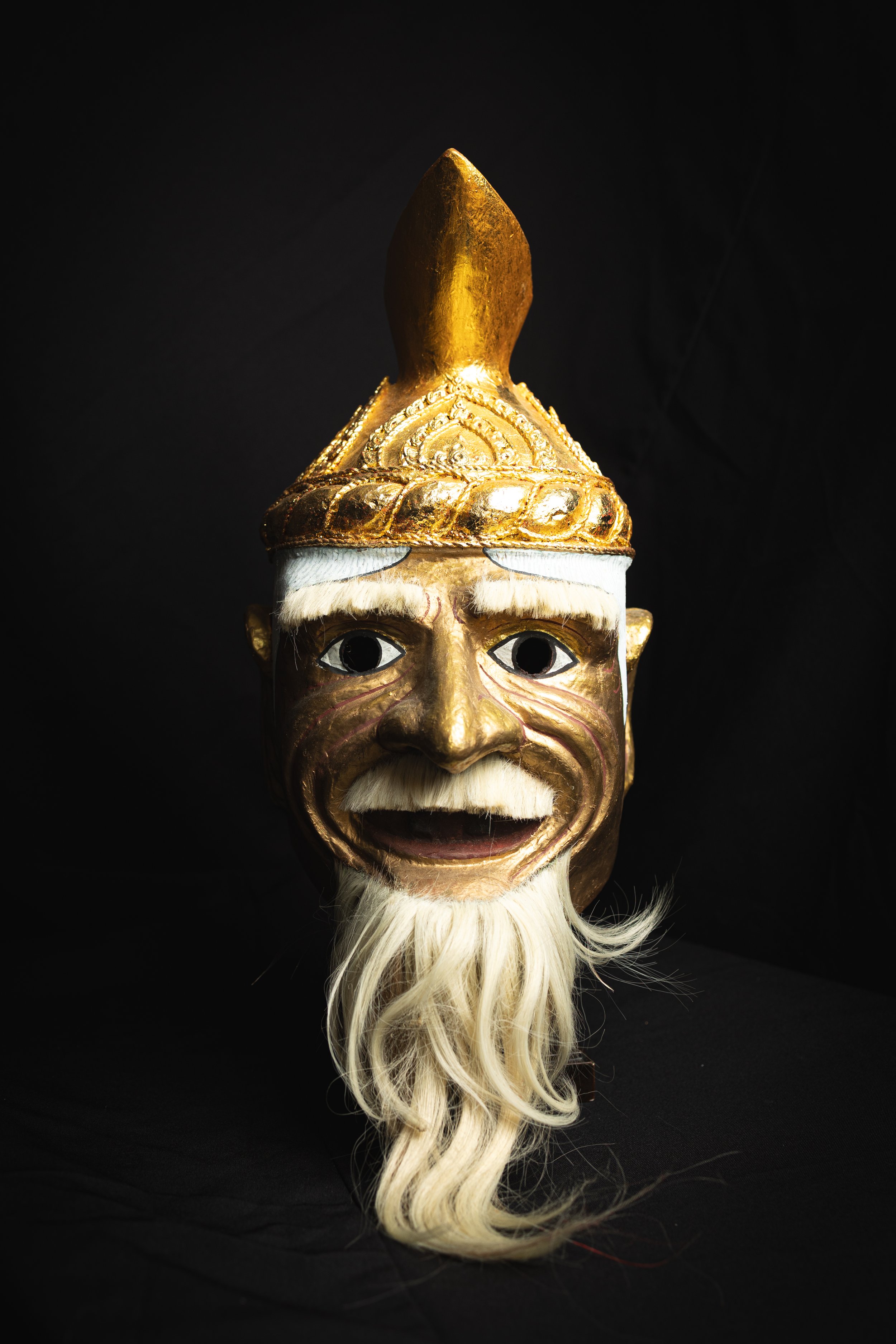 A decorative mask with a gold and white face, intricate patterns, white eyebrows, and a long white beard, set against a black background.