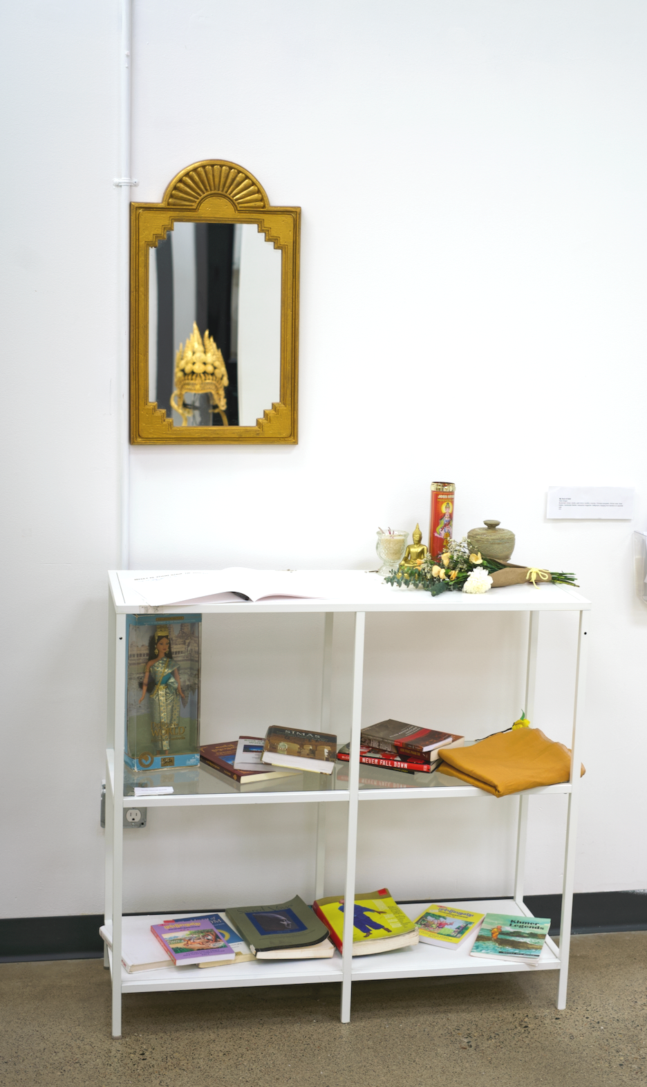 A white shelf with books and religious items, including a small Buddha statue, a candle, and flowers. Above the shelf, a mirror with a decorative gold frame reflects a golden temple ornament.