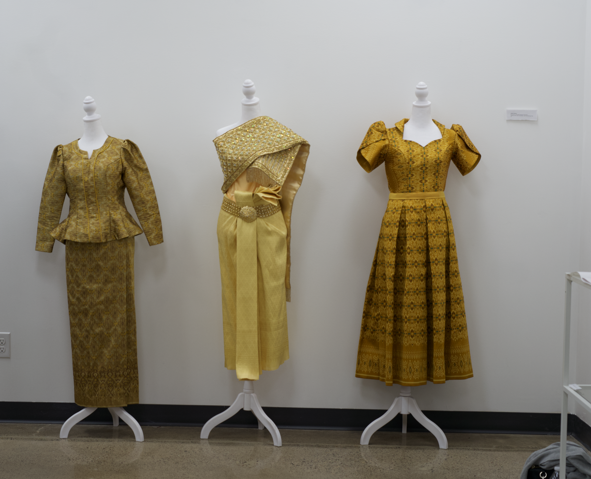 Three mannequins dressed in traditional gold and yellow garments, displayed against a plain white wall in an exhibition setting.