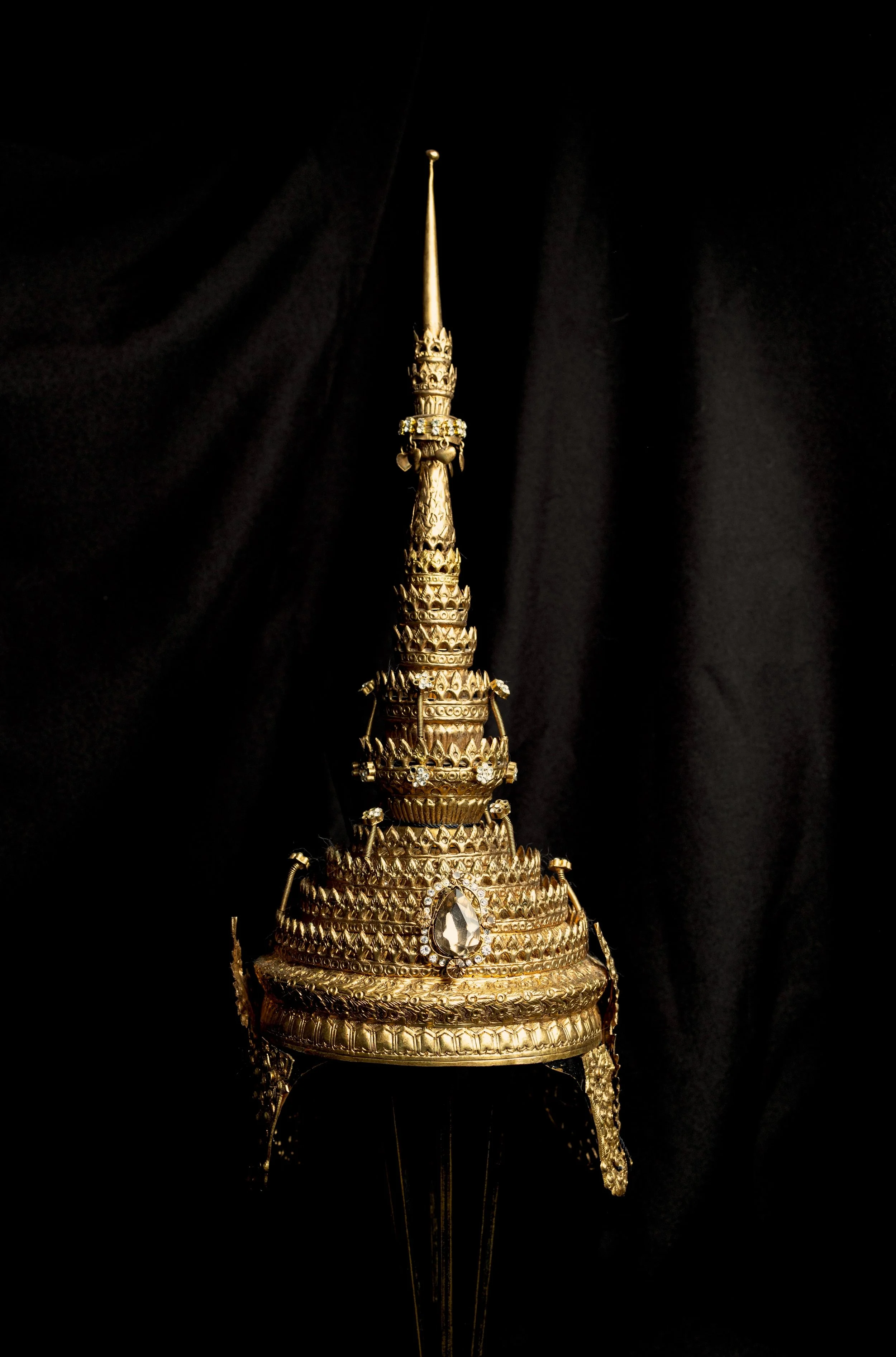 A tall, ornate golden ceremonial headdress with intricate designs and embellishments, including a large gemstone, set against a black background.
