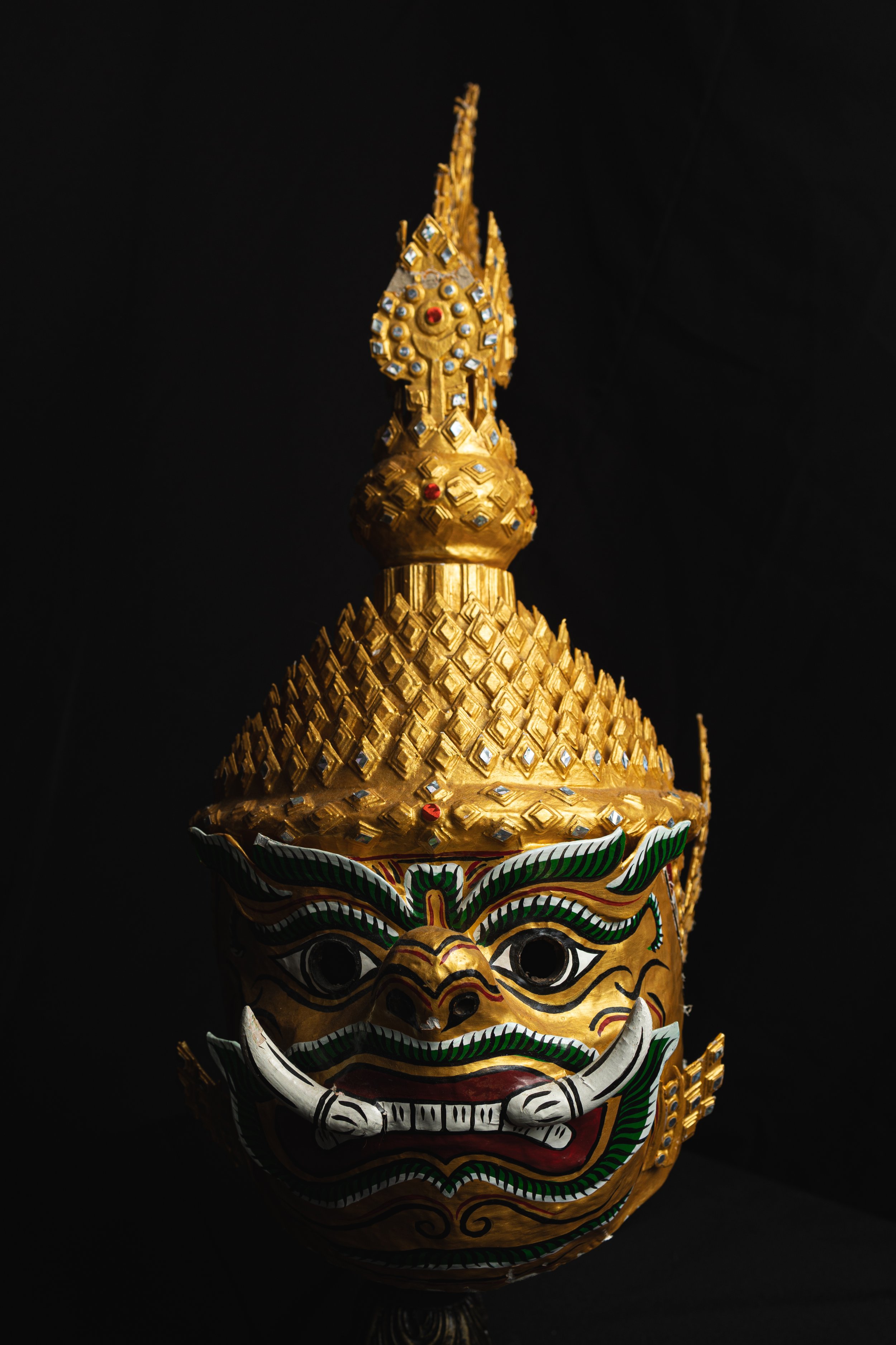 A detailed gold and painted mask of a mythological creature with a fierce expression, large eyes, and ornate decorations, set against a black background.