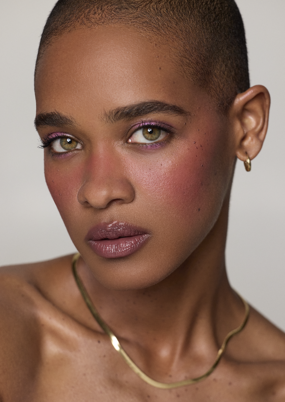 Close-up portrait of a woman with short dark hair, gold hoop earrings, and a gold necklace. She has light green eyes, pink eyeshadow, and glossy lips, with a natural glow and freckles on her face.