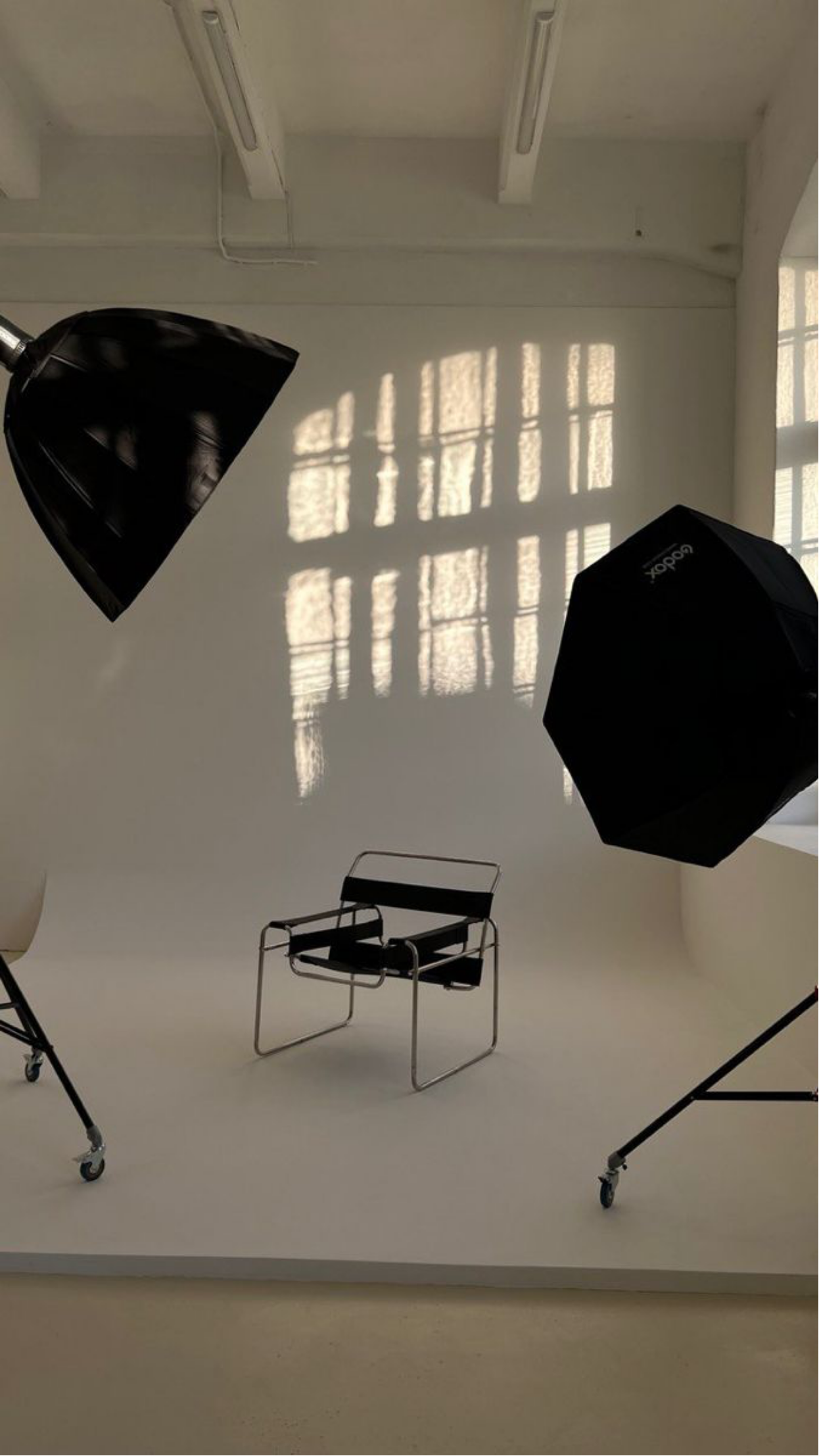 A photography studio with two studio lights on stands, a black and chrome chair in a white room with the sunlight casting shadow patterns through windows.