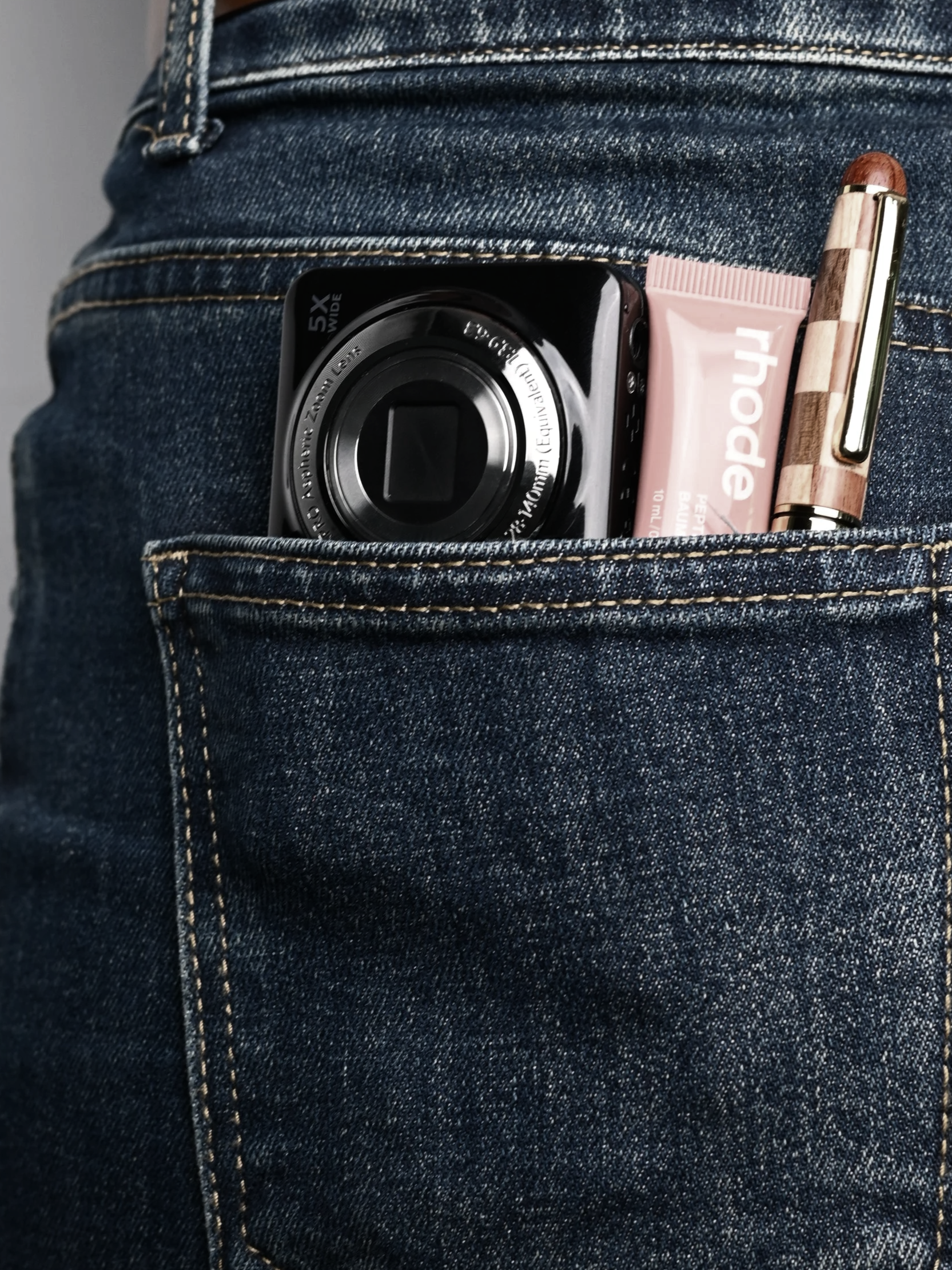 Close-up of a person wearing blue denim jeans with a pocket. Inside the pocket, there is a small black camera, a pink tube of lotion, and a wooden and gold pen.
