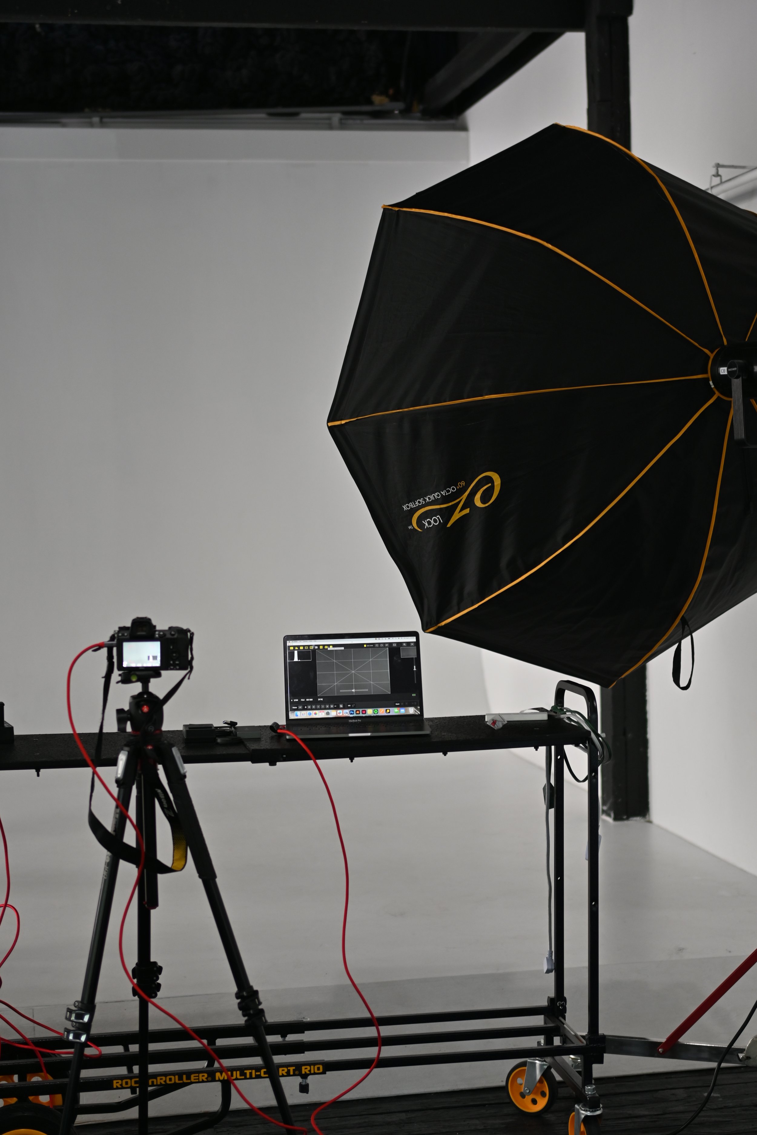 A photography setup with a camera on a tripod, a computer monitor, and a large black umbrella with orange trim on a stand in a studio with a plain white backdrop.