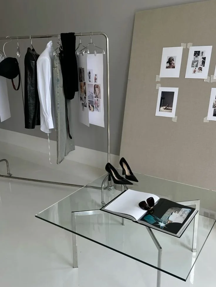 Fashion design workspace with clothing rack holding various garments, high heels on a glass table, and photo references taped to a beige wall.