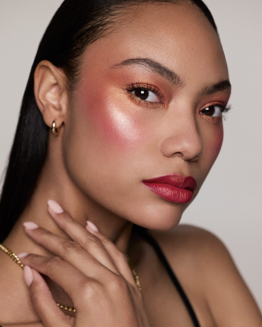 A woman with dark hair, wearing gold hoop earrings, and makeup that includes reddish eyeshadow, blush, and red lipstick, posing with her hand near her face.