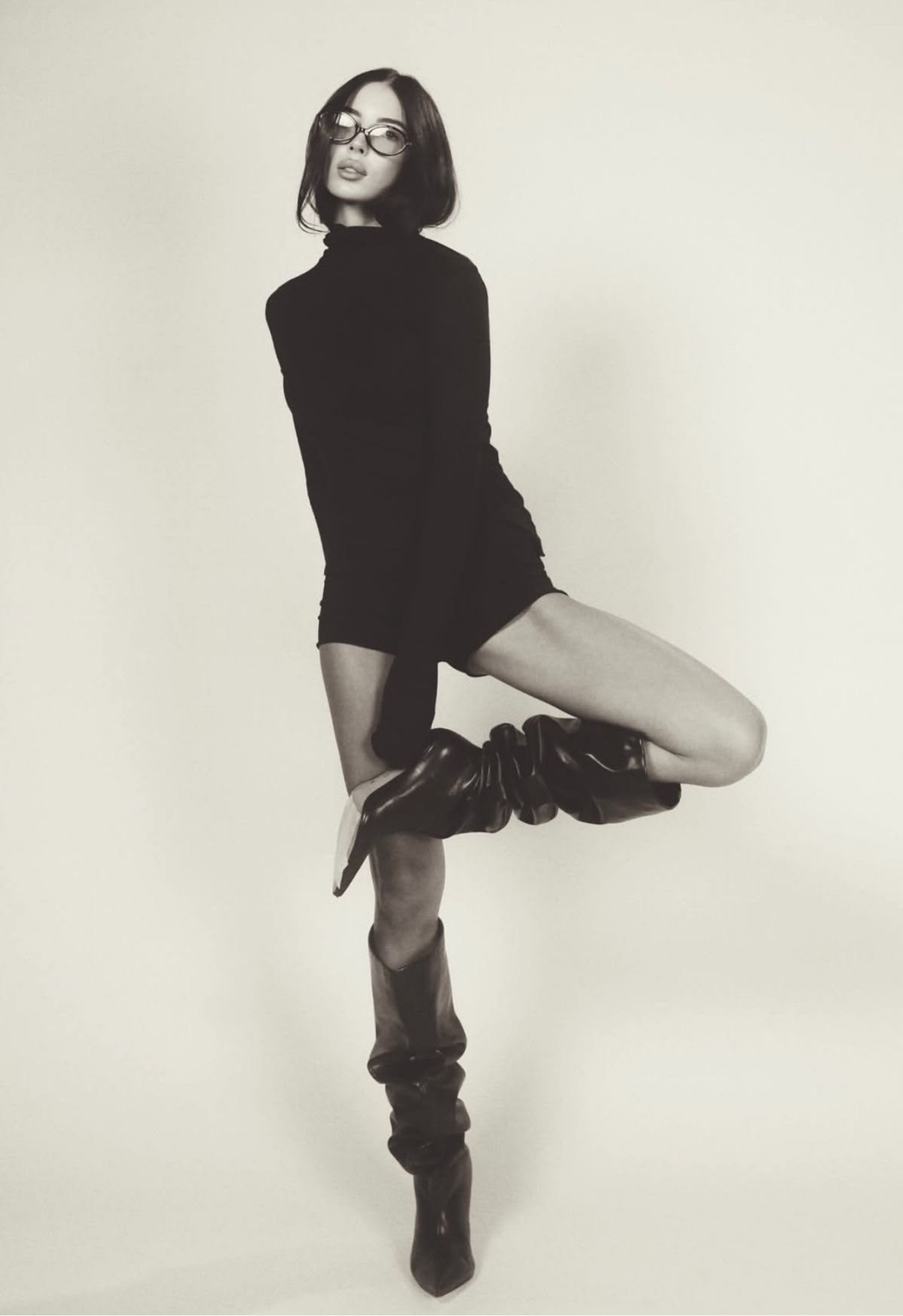 A woman with short dark hair wearing glasses, a black dress, and knee-high boots, striking a fashion pose against a plain light background.