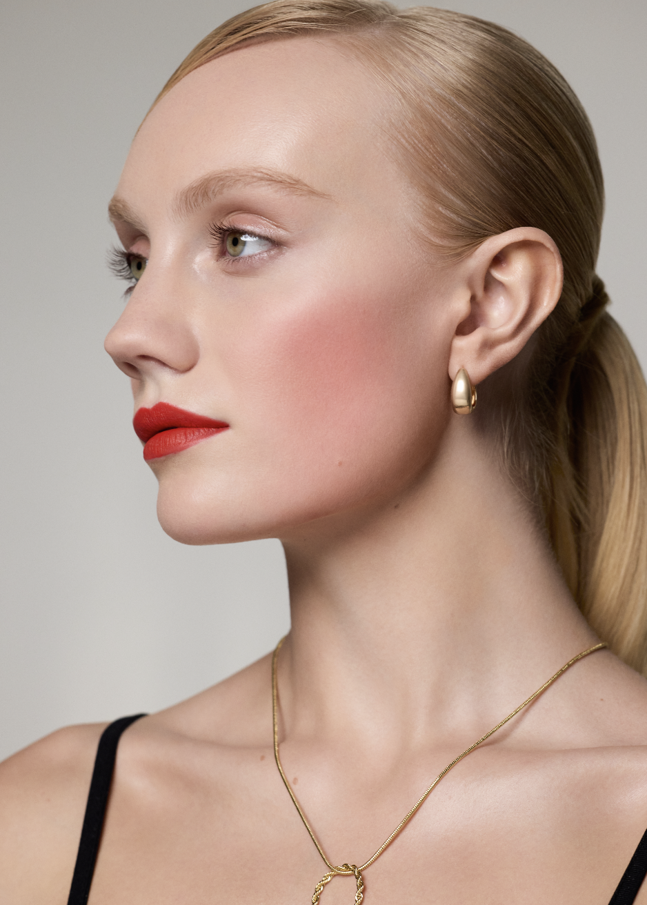 Close-up profile of a young woman with blonde hair, red lipstick, gold earrings, and a gold necklace, looking to the left.