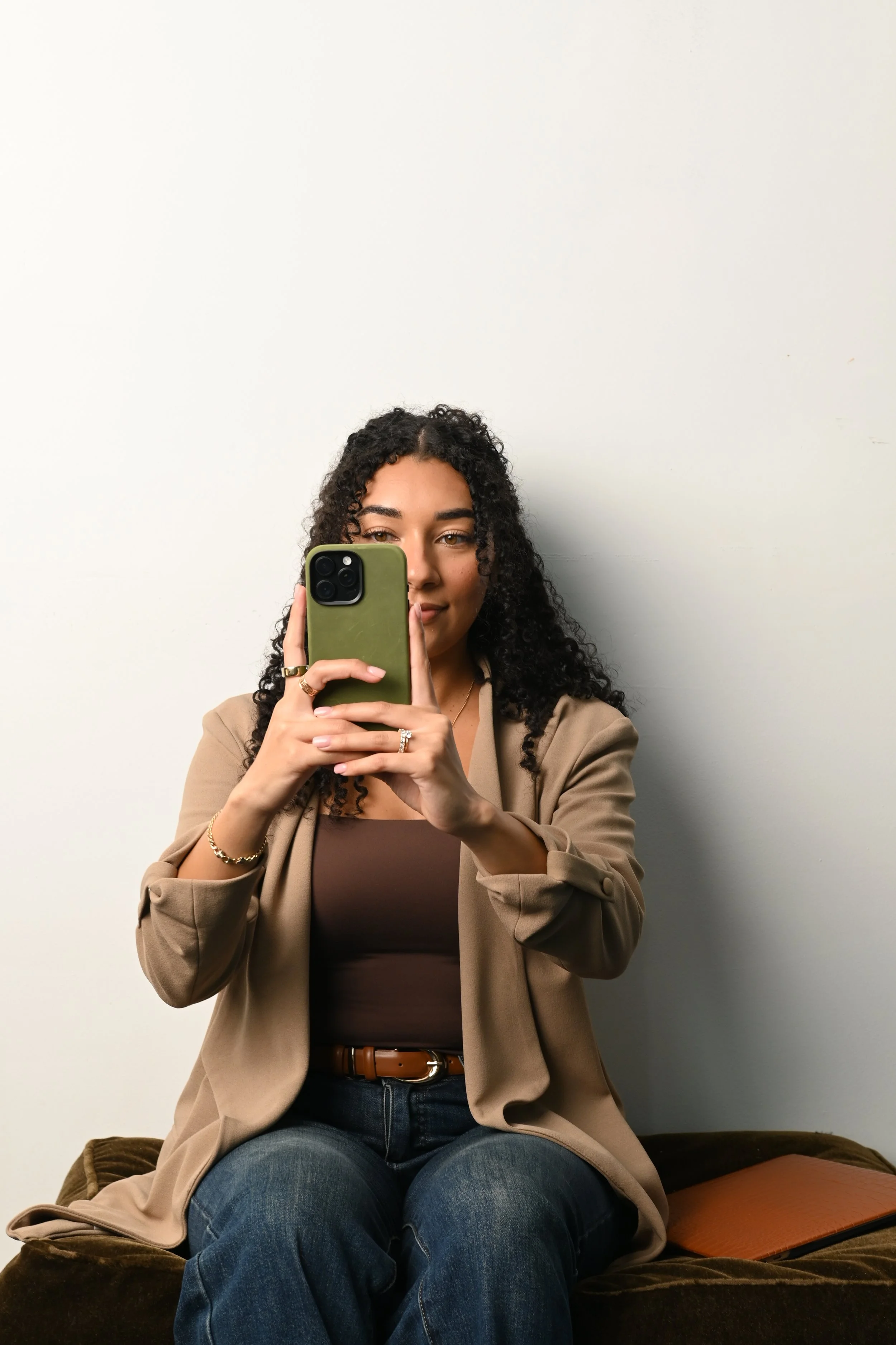 A woman with curly dark hair taking a selfie with a smartphone while sitting on a brown couch against a plain white wall, wearing a beige blazer over a brown top and blue jeans.