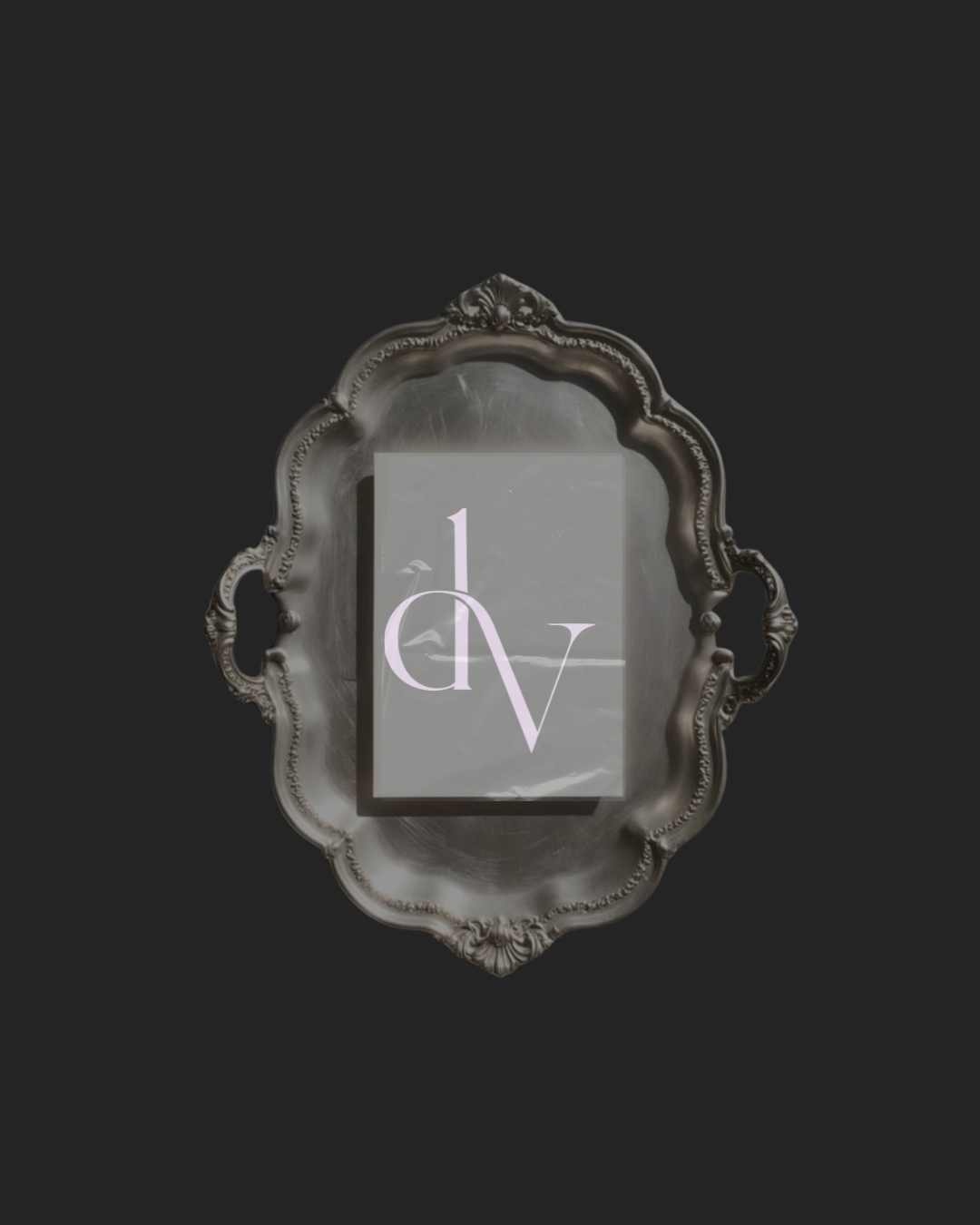 An ornate silver tray with a decorative border and handles, placed on a dark background. On the tray, there is a rectangular card or sign with the letters 'd' and 'v' and the number '1' in a stylized font.