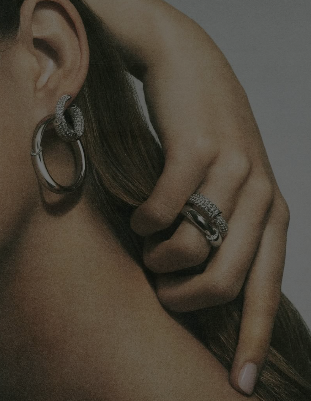 Close-up of a person's ear with multiple earrings and hand with rings, touching their hair and shoulder.