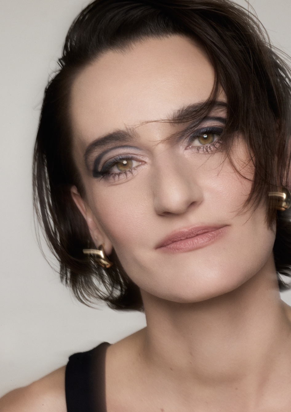 A close-up portrait of a woman with short dark hair, wearing makeup with bold eyeshadow, and a black top. She has gold earrings, and the background is plain and light-colored.