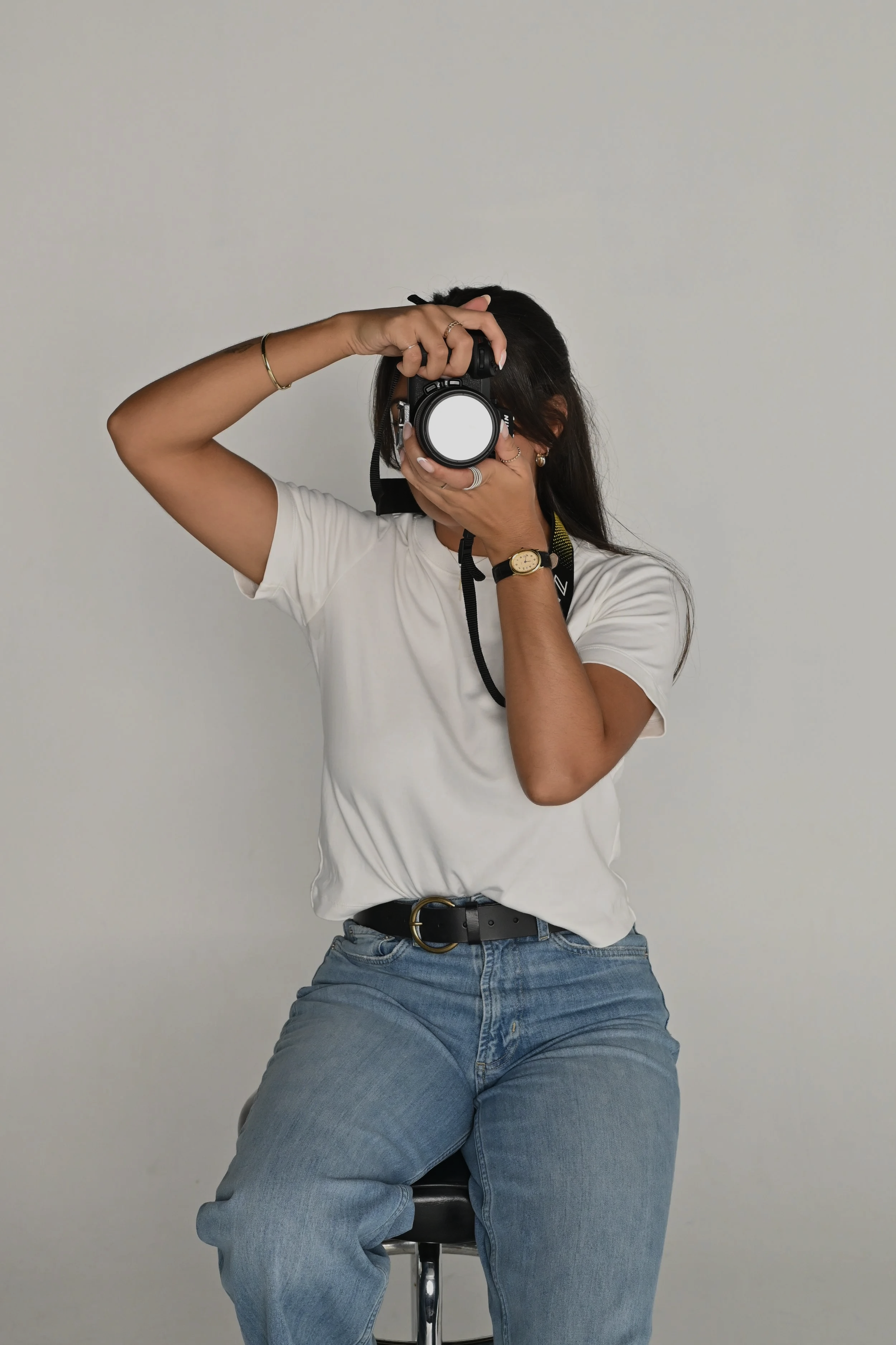 A woman sitting on a stool taking a mirror selfie with a DSLR camera, wearing a white t-shirt, blue jeans, and gold jewelry, against a plain white wall.