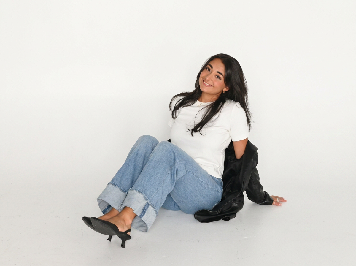 A woman with long dark hair, wearing a white t-shirt, light blue jeans with cuffs, and black high heels, is sitting on the floor against a white background. She is smiling and looking at the camera.