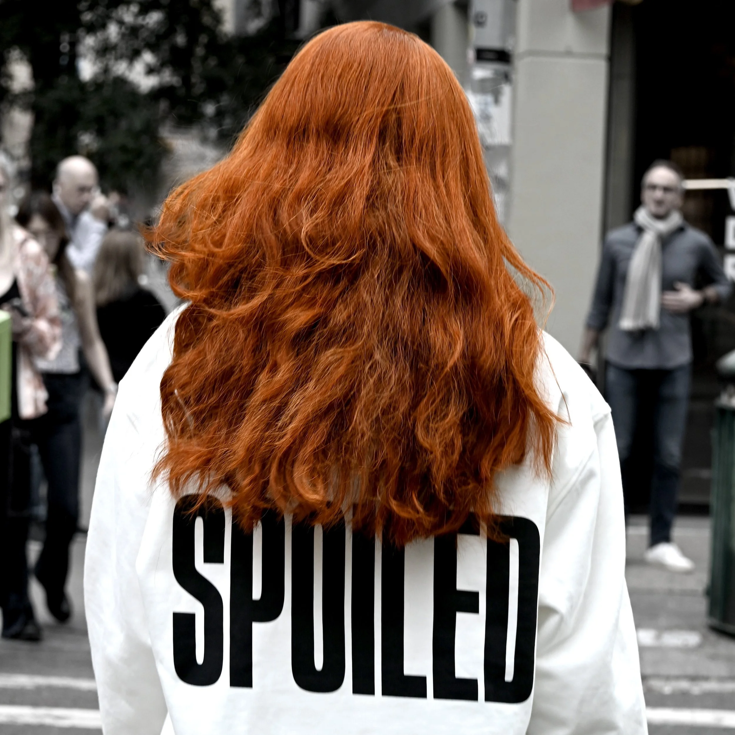A person with long, curly red hair standing behind a glass door with the word 'SOCIETY' written on it. The person's face is not visible, and there are blurred people in the background.