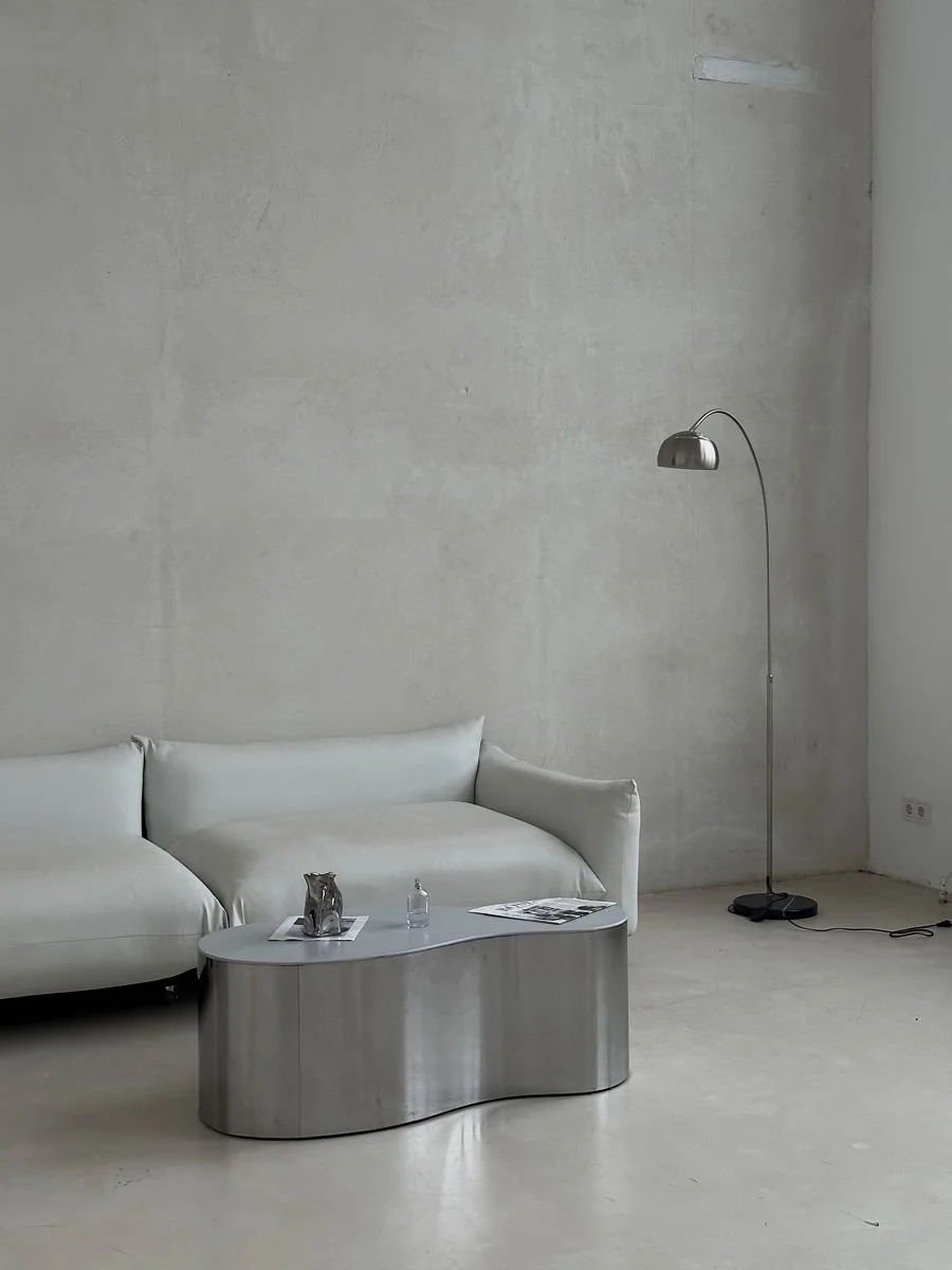 Minimalist living room with a white sofa, a metallic coffee table, a modern floor lamp, and some magazines and a bottle on the table.