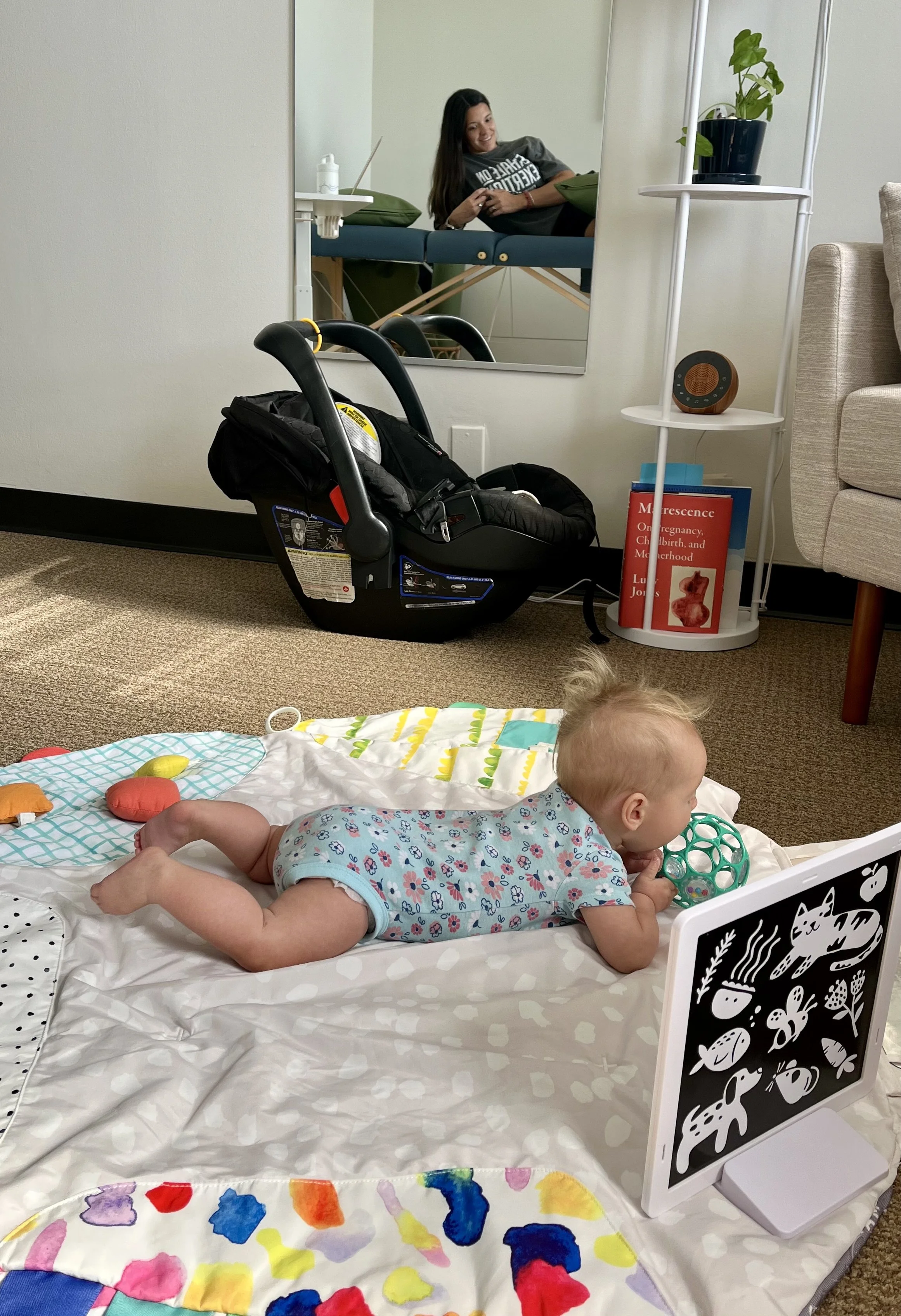 A baby plays on a colorful playmat while her mother receives physical therapy treatment during a baby and me session