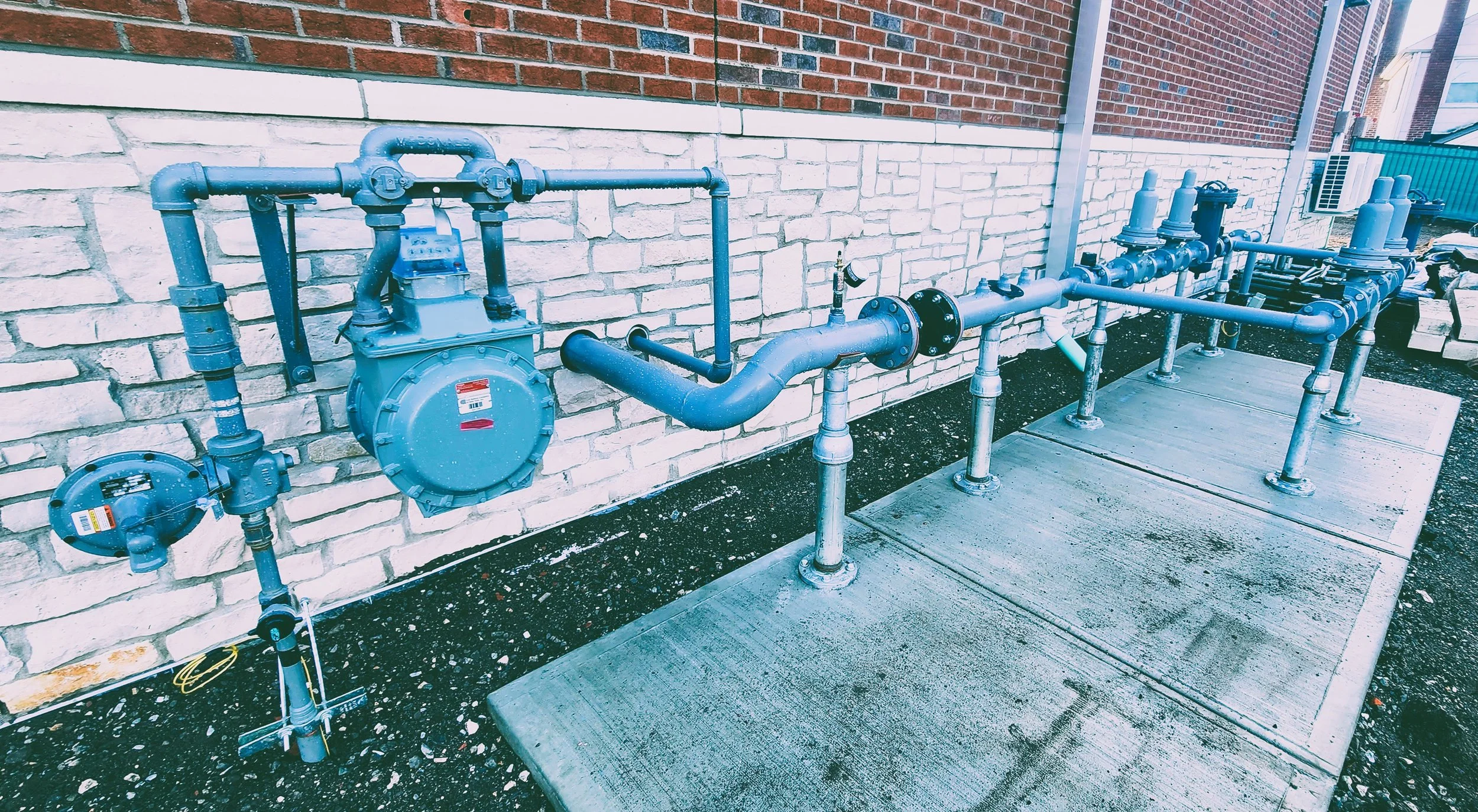 Exterior view of plumbing and gas meters attached to a brick wall, with metal pipes running along the wall and a concrete platform on the ground.