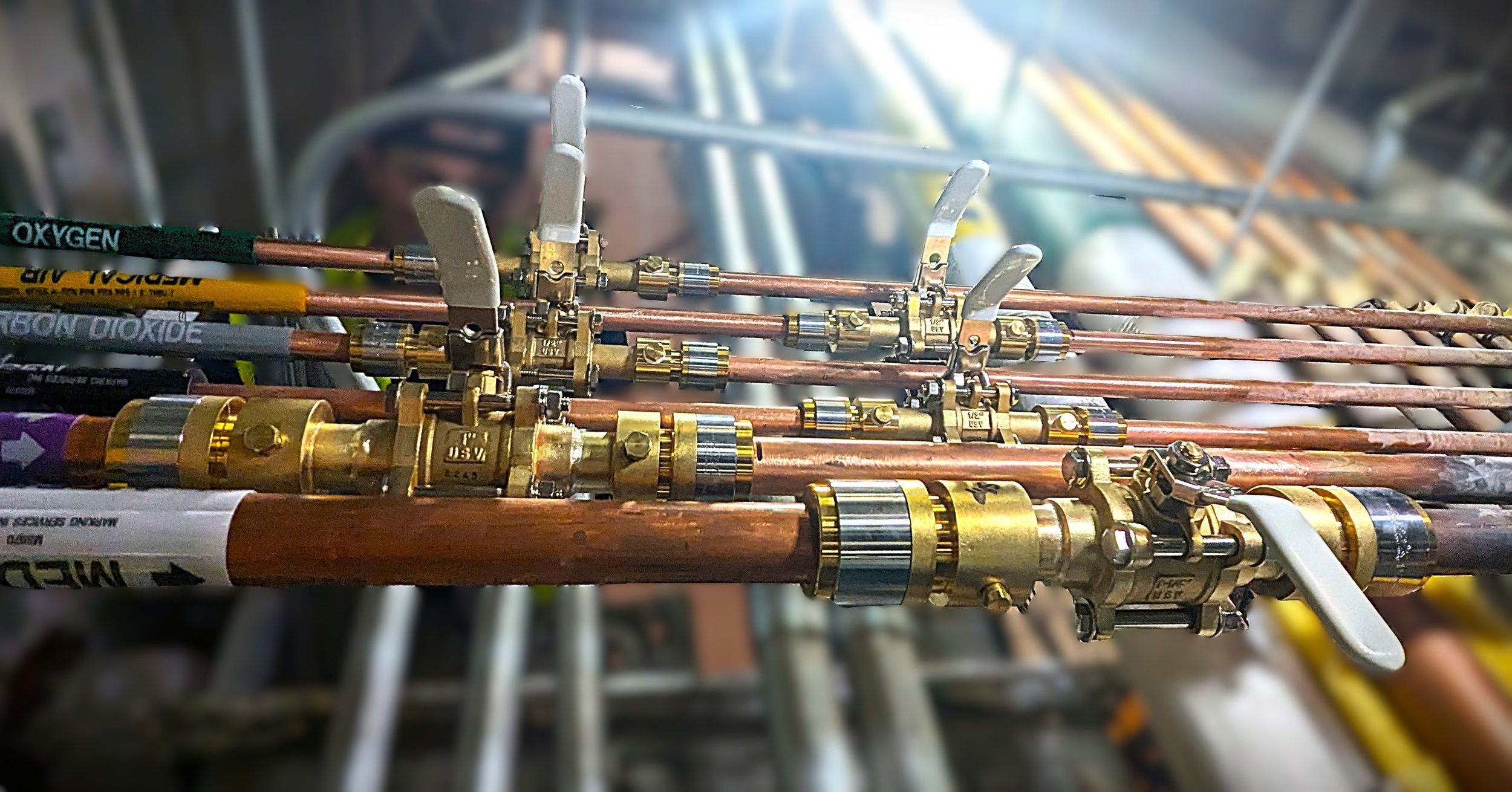 Close-up of several brass and copper pipes with valves, some labeled with oxygen and other gases, likely part of industrial or medical equipment.