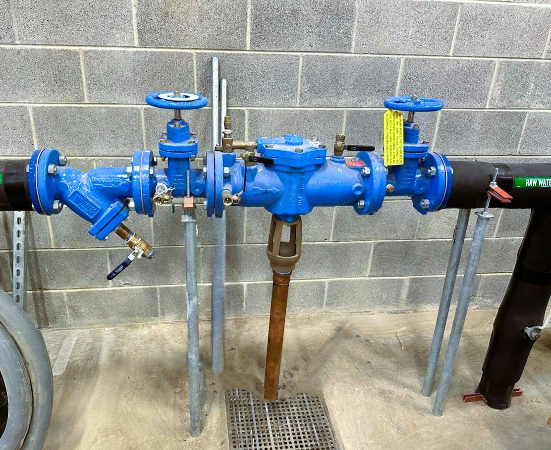 Blue water pipe with valves, connected to a black pipe, mounted on metal supports against a gray cinder block wall.
