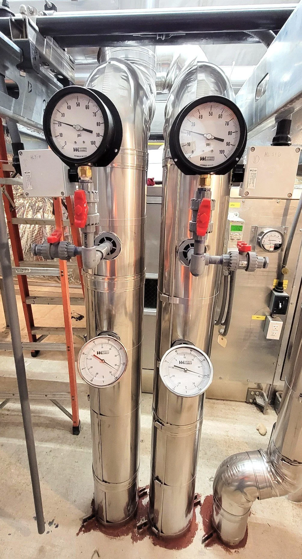 Two stainless steel vertical tanks with pressure gauges and valves, connected to pipes in an industrial setting.