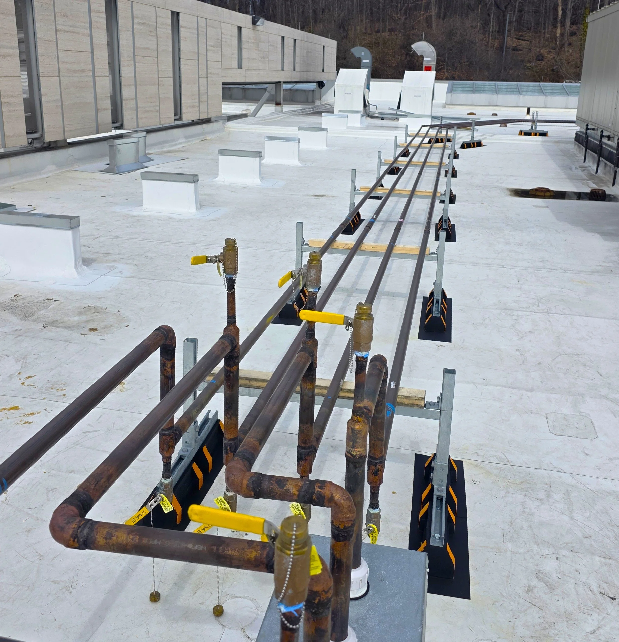 Construction area on the roof of a building under development, showing various metal pipes, vents, and fixtures, with an exterior wall on the left and a background of trees and hillside.