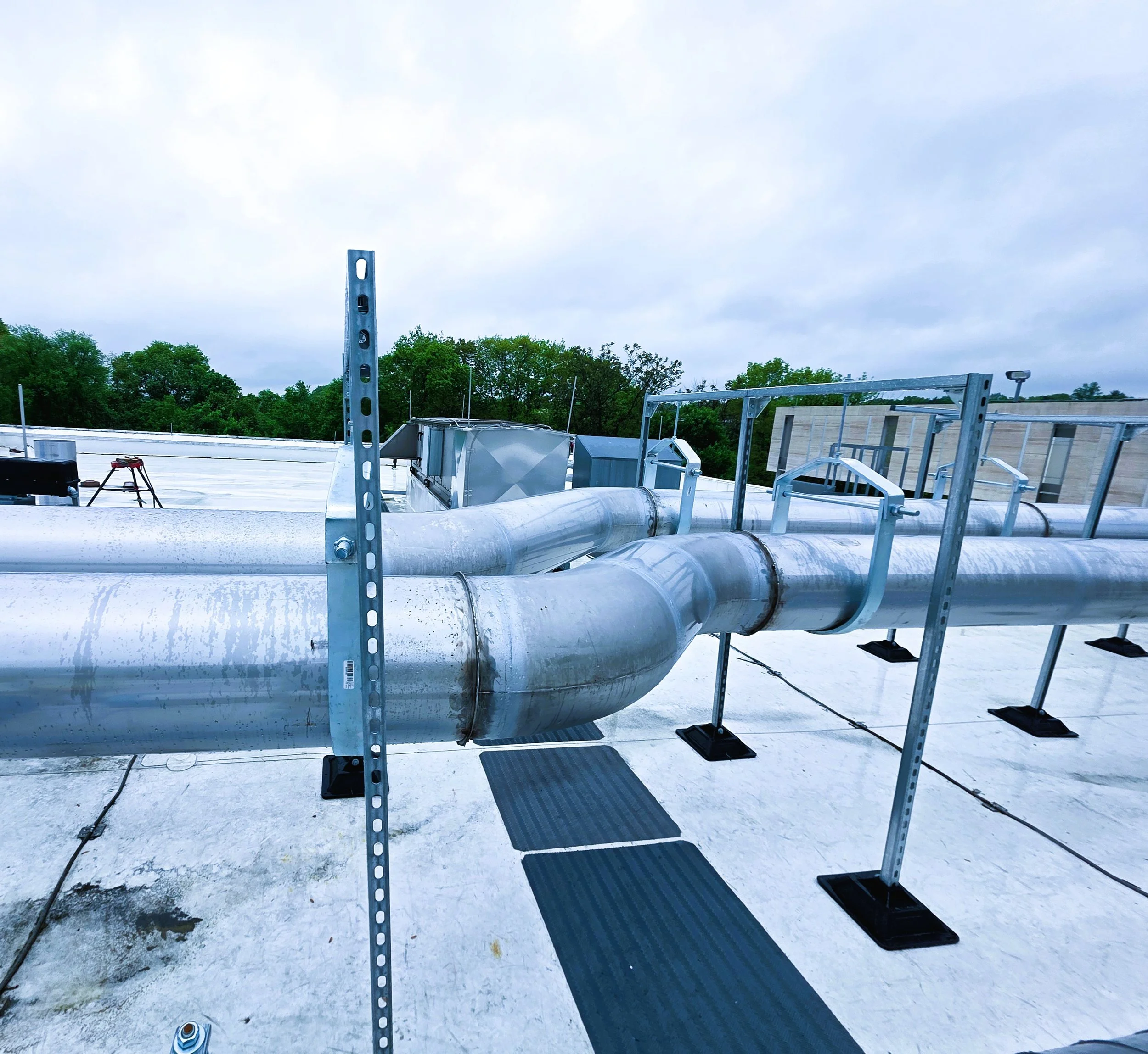 View of a building rooftop with metal pipes, ductwork, and structural supports with trees and a cloudy sky in the background.