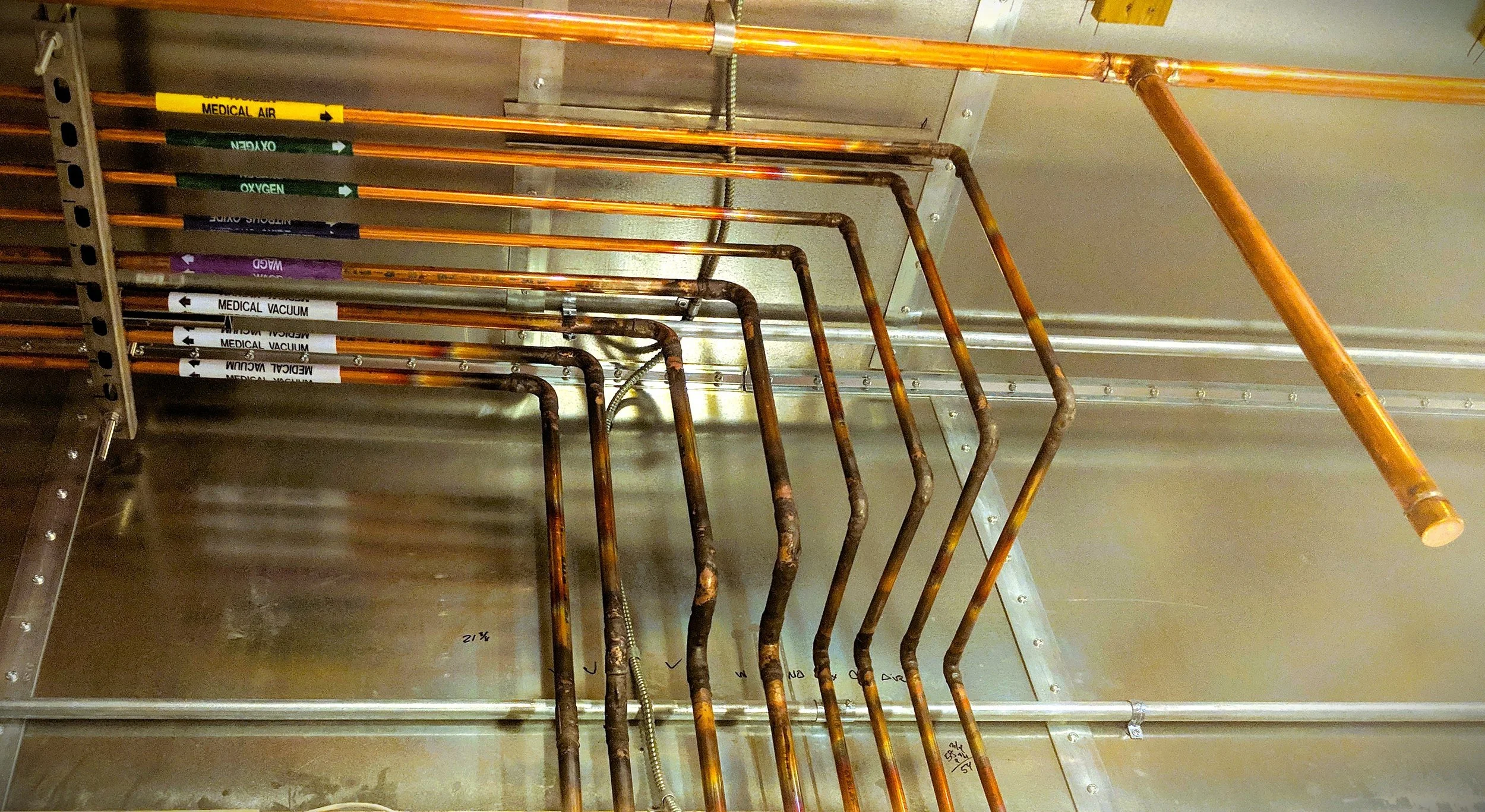 A view inside an industrial or utility cabinet showing several copper pipes labeled with various medical gases such as 'Medical Air,' 'Oxygen,' 'Vacuum,' and 'Wall Suction.' The pipes are arranged parallel and connected to a metallic structure with l