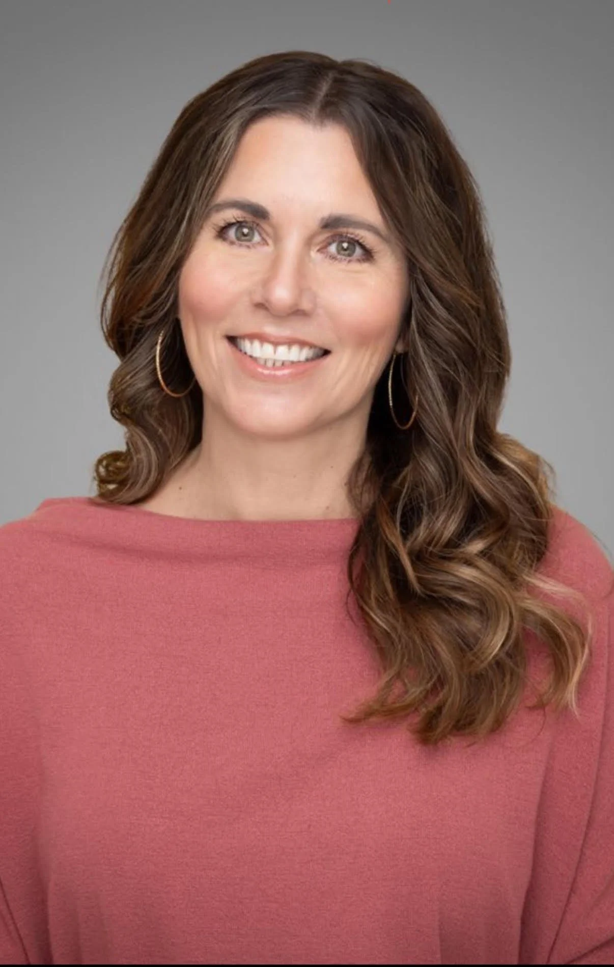 A woman with long wavy brown hair, light skin, wearing a pink sweater and hoop earrings, smiling against a plain gray background.
