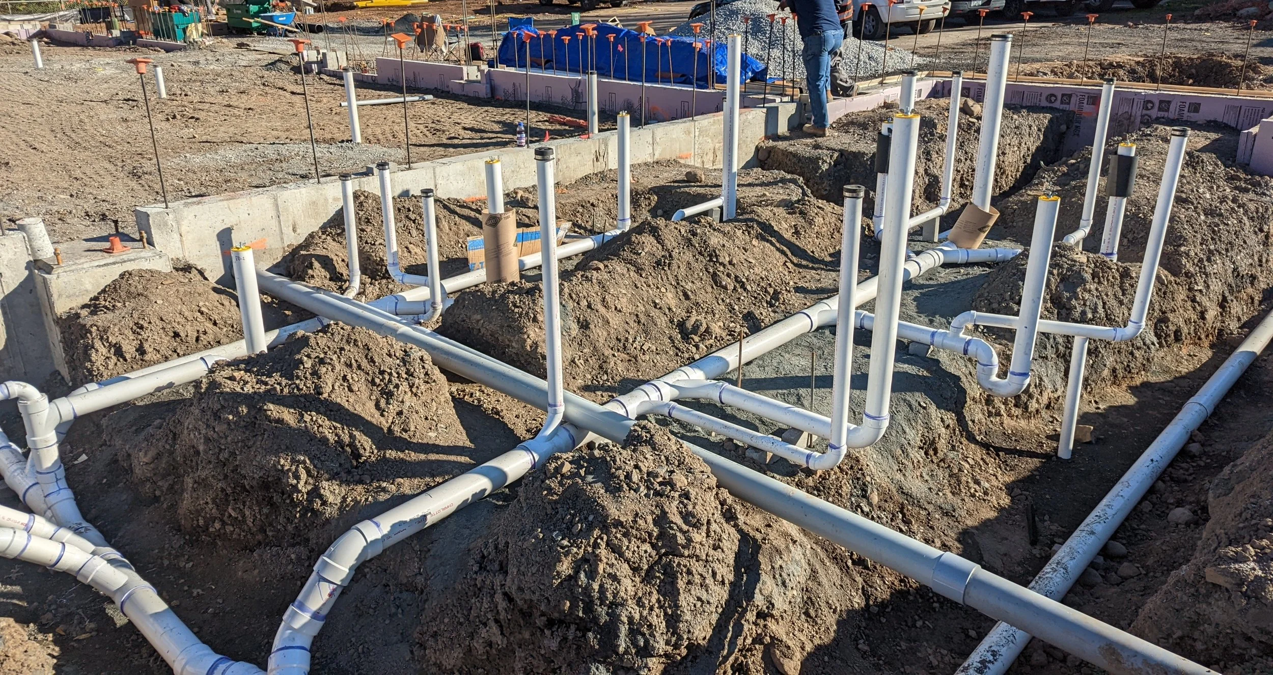 Construction site with exposed white PVC pipes laid out in trenches for plumbing, surrounded by dirt and construction materials.
