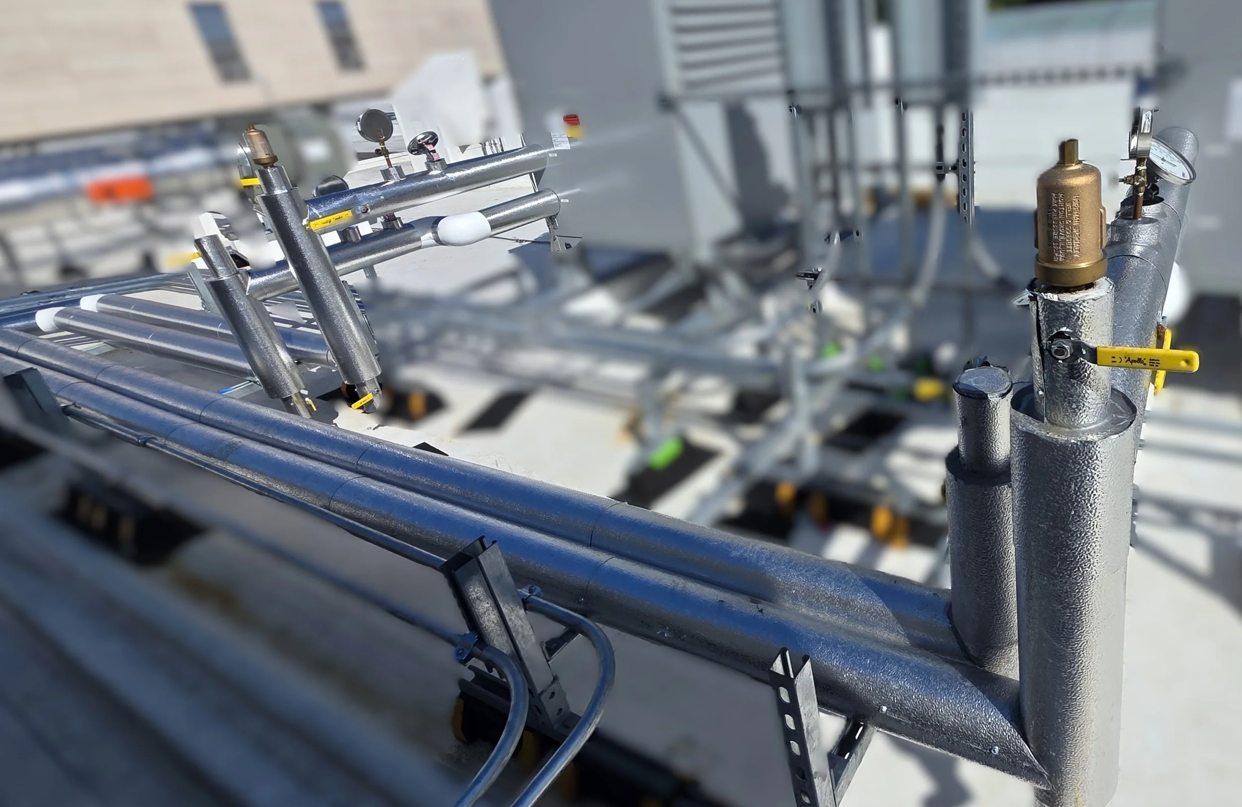 Close-up of metallic industrial pipes and valves on a rooftop, with a building in the background.