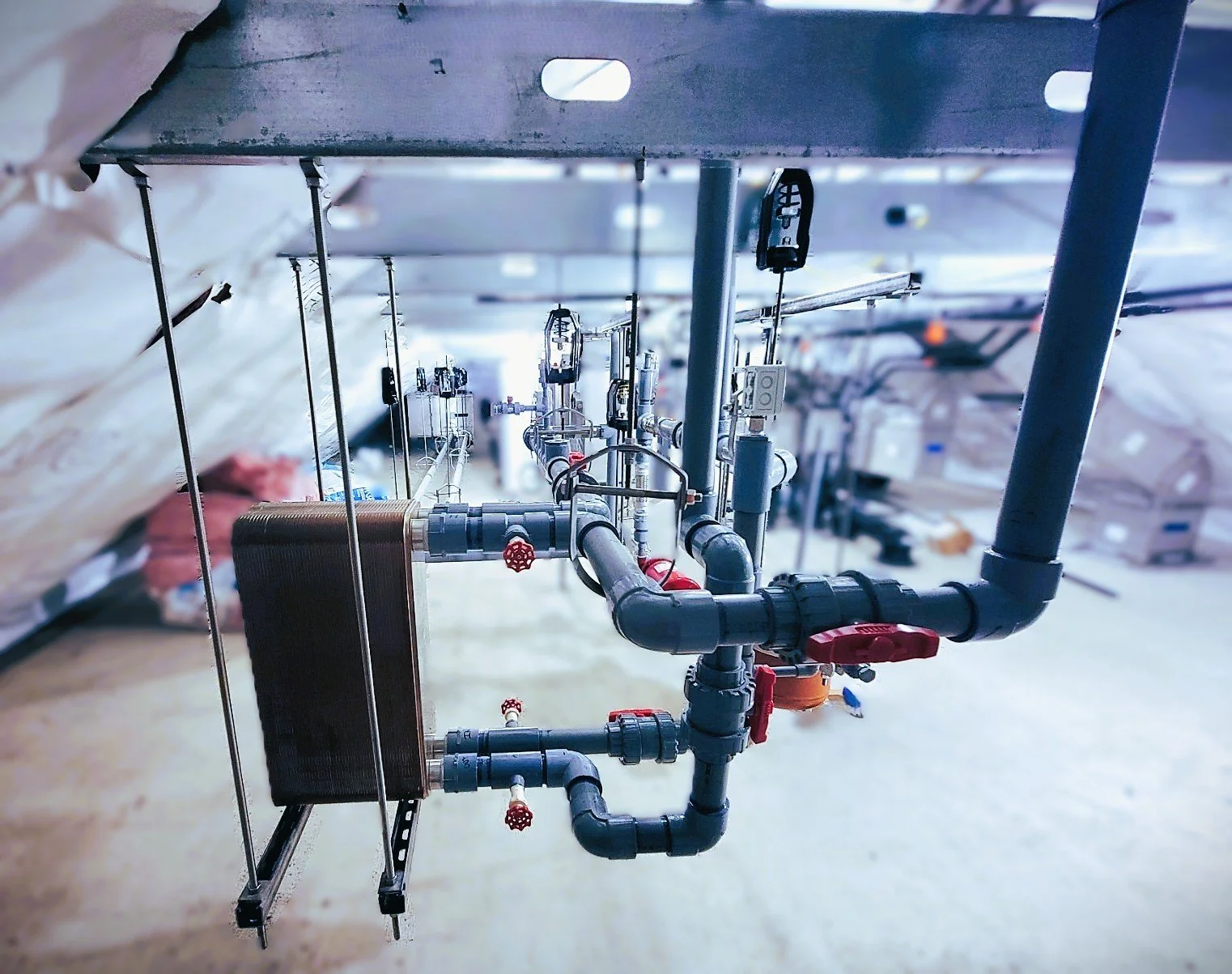 A network of black and gray pipes, valves, and gauges on a ceiling in an industrial or mechanical setting.